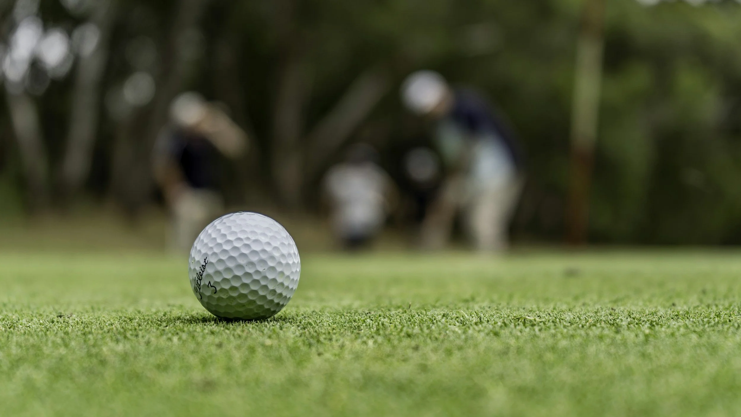 Une balle de golf blanche posée sur l'herbe, avec deux personnes portant des casques de golf en arrière-plan, floues.