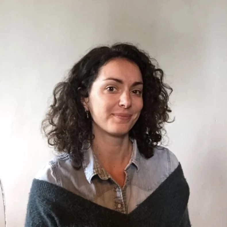 Une femme avec des cheveux bouclés, portant une chemise et un pull, souriant légèrement, posant contre un mur blanc.