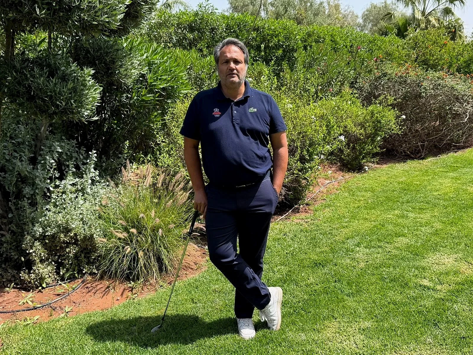 Un homme debout dans un jardin, tenant un bâton de golf, portant une chemise bleue Lacoste et des pantalons noirs, entouré de plantes et d'arbustes.