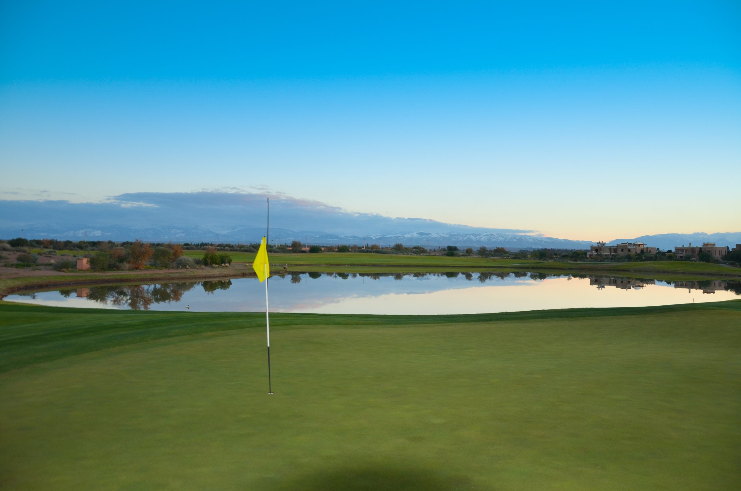 Un green de golf avec un drapeau jaune devant un lac, avec un paysage de montagnes et des bâtiments résidentiels en arrière-plan, sous un ciel bleu clair.