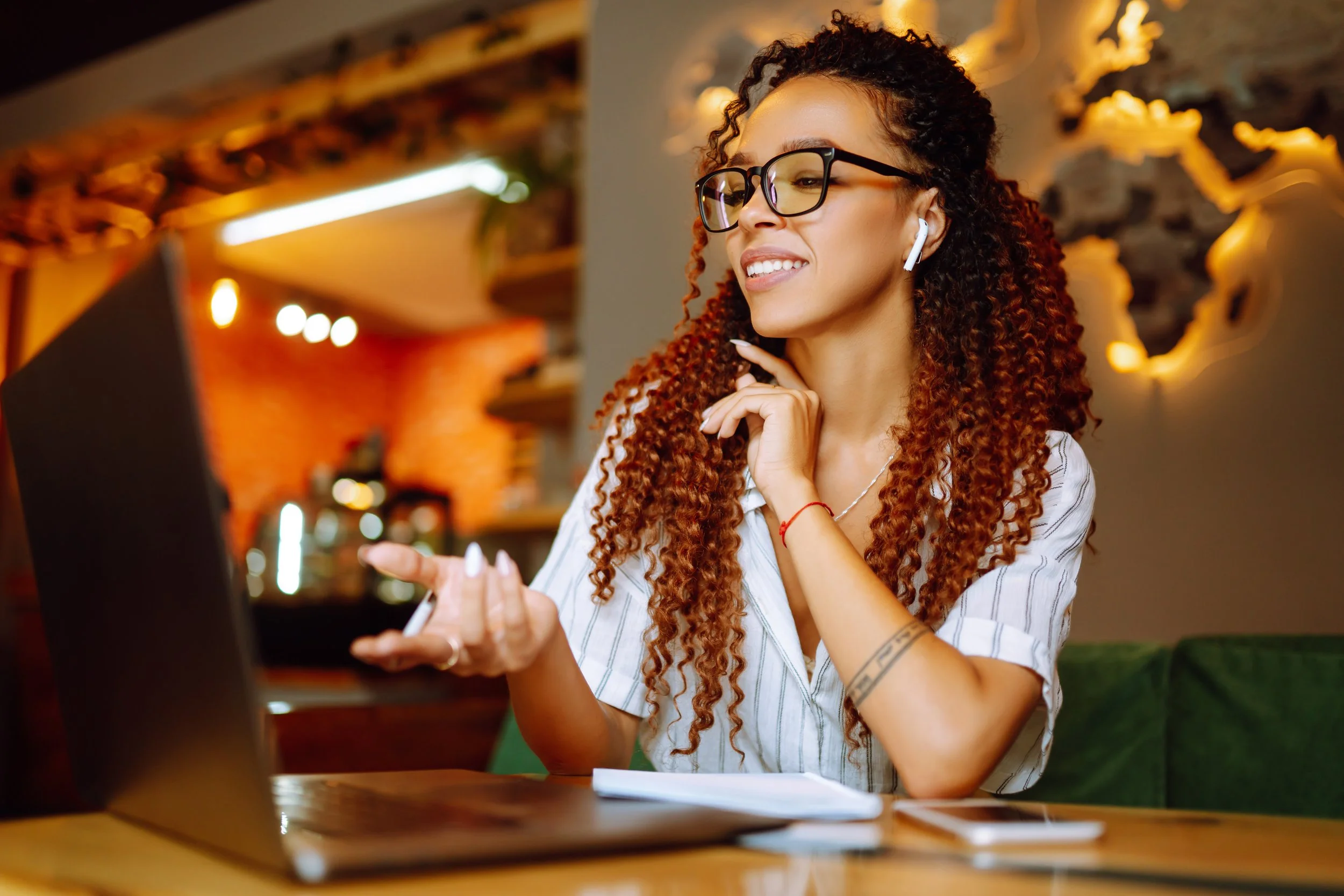 A young woman with curly hair, glasses, and earbuds, smiling and engaging in an one-on-one online training for her bookkeeping client.
