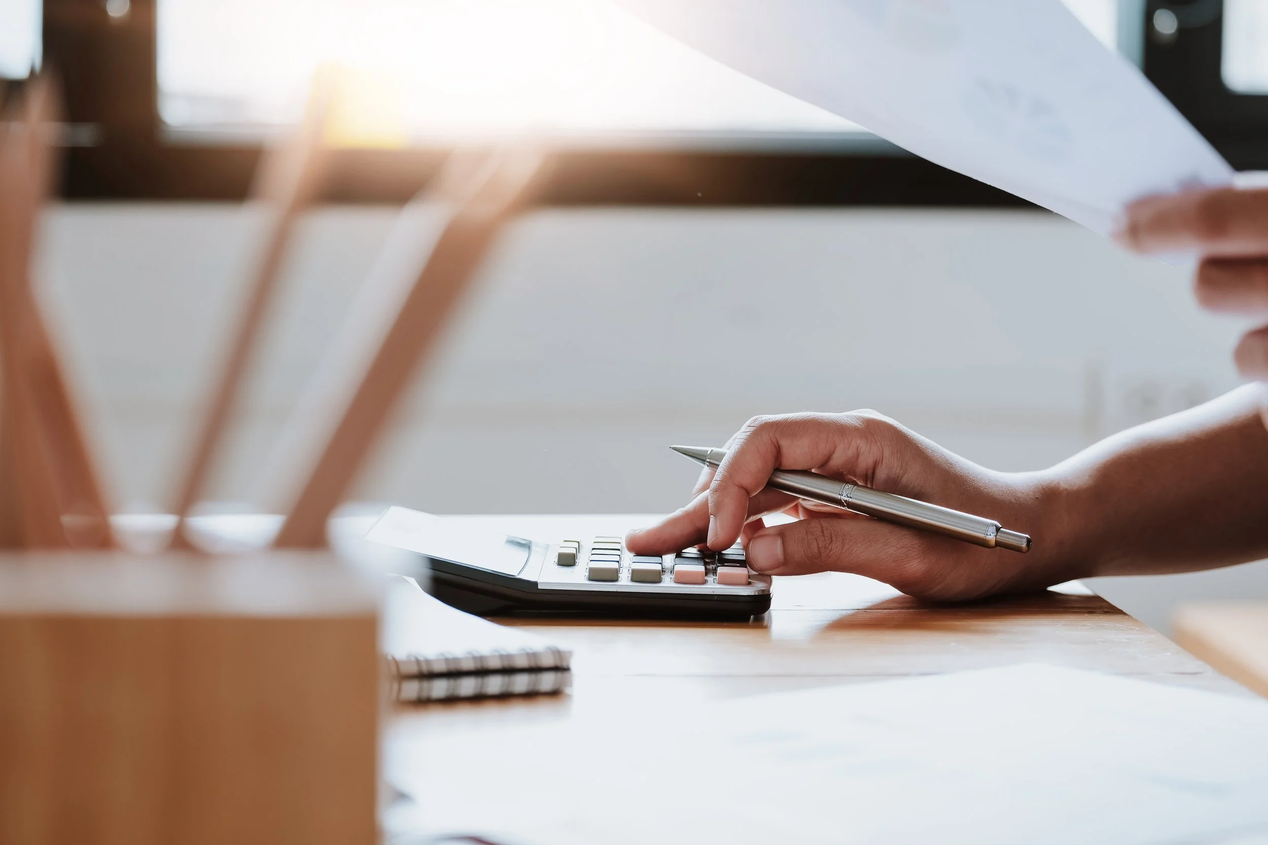Person using a calculator while holding a pen and paper, at a desk with office supplies.