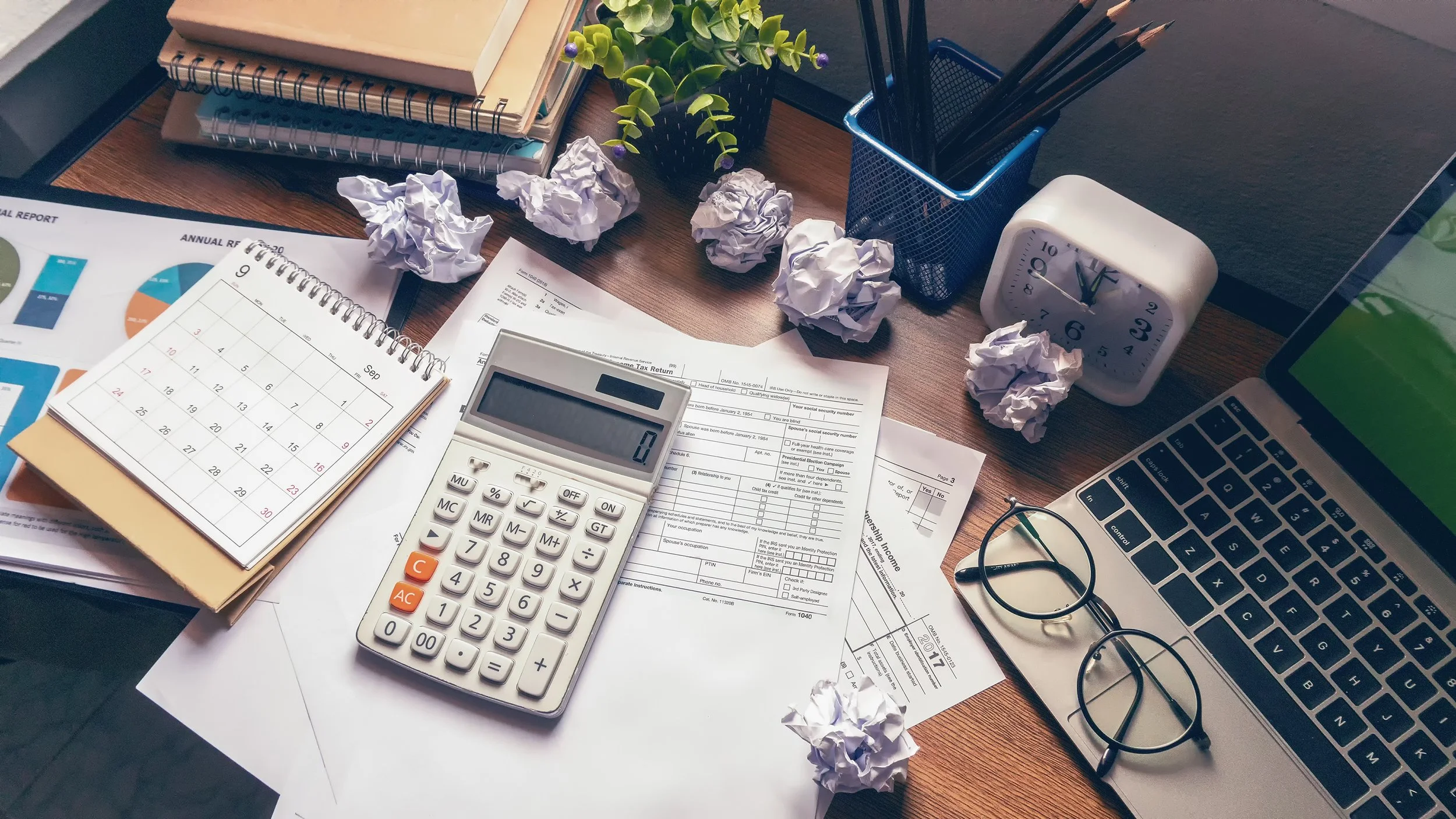 A cluttered desk with crumpled papers, a calendar, a calculator, a pair of glasses, notebooks, a clock, some pens, a laptop, and a potted plant.