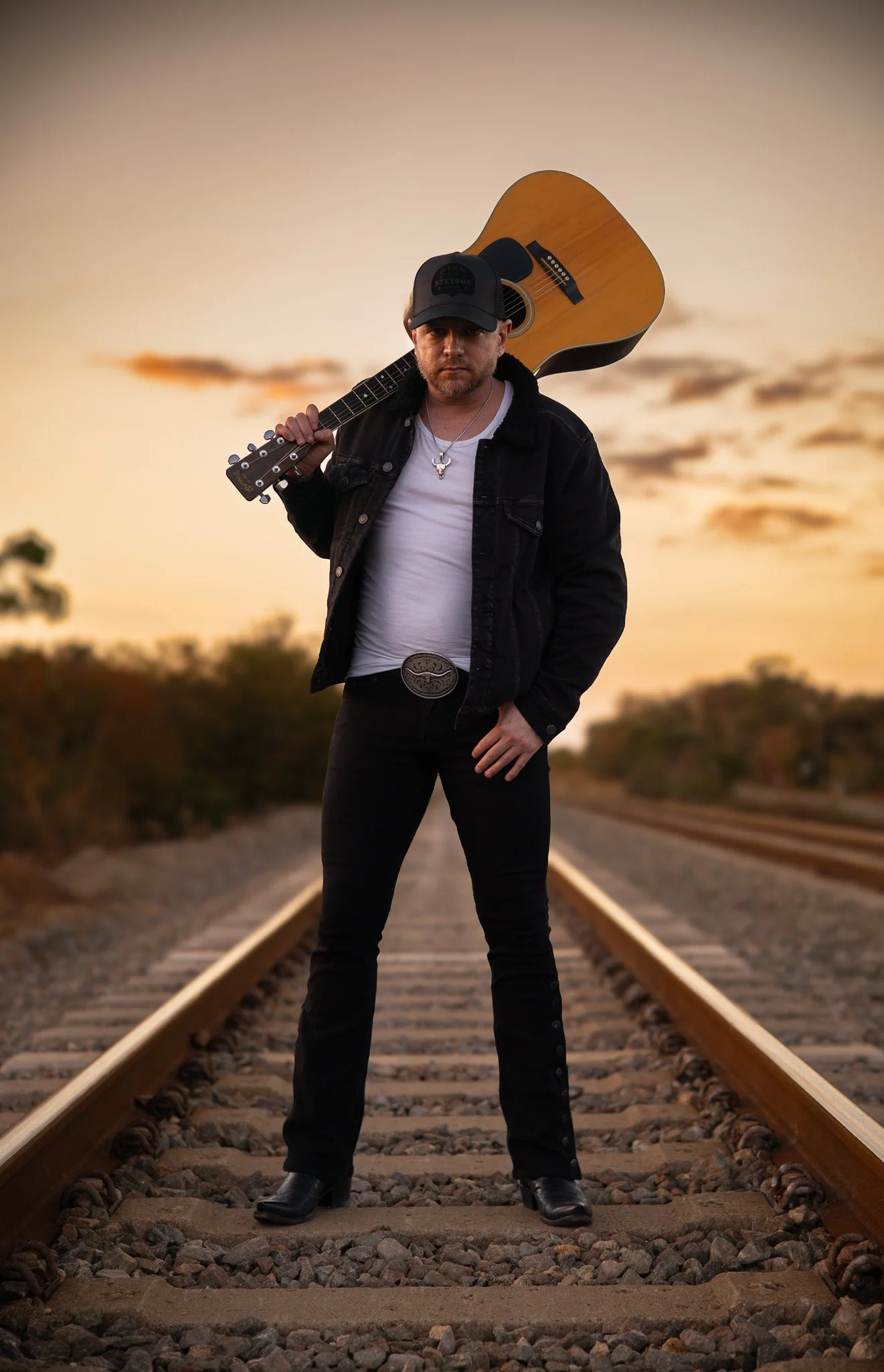 A man standing on railroad tracks during sunset, holding an acoustic guitar over his shoulder, wearing a black jacket, white shirt, black jeans, and a black cap.