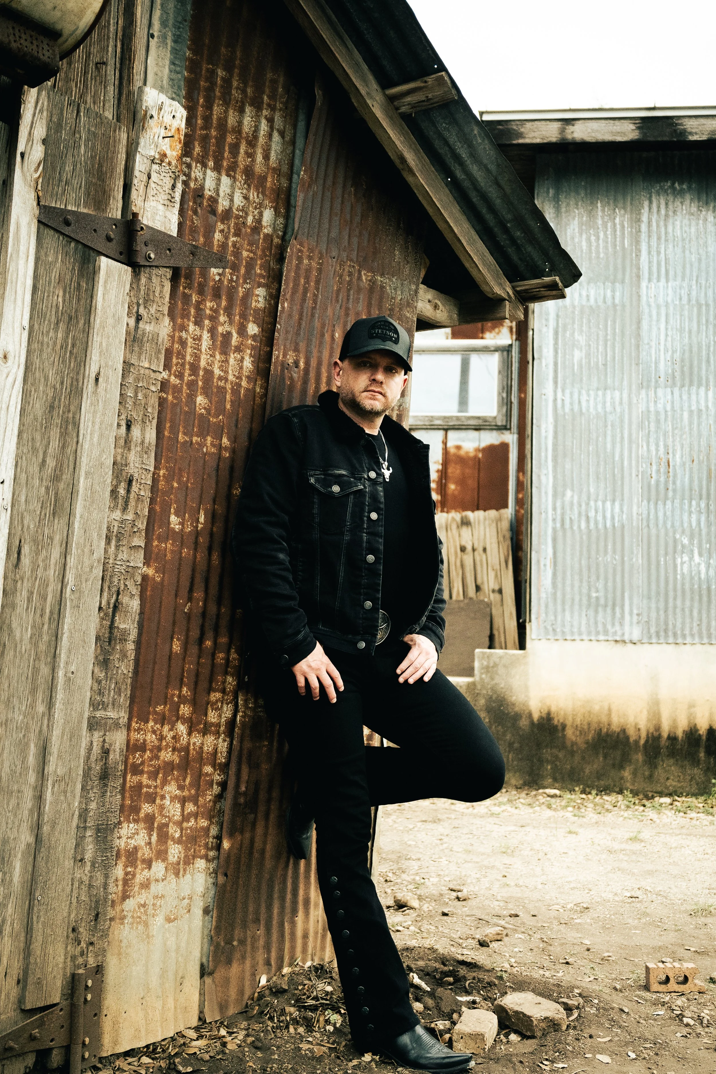 A man in a black denim jacket and black pants leaning against a weathered, rusty metal wall of a rustic shed.