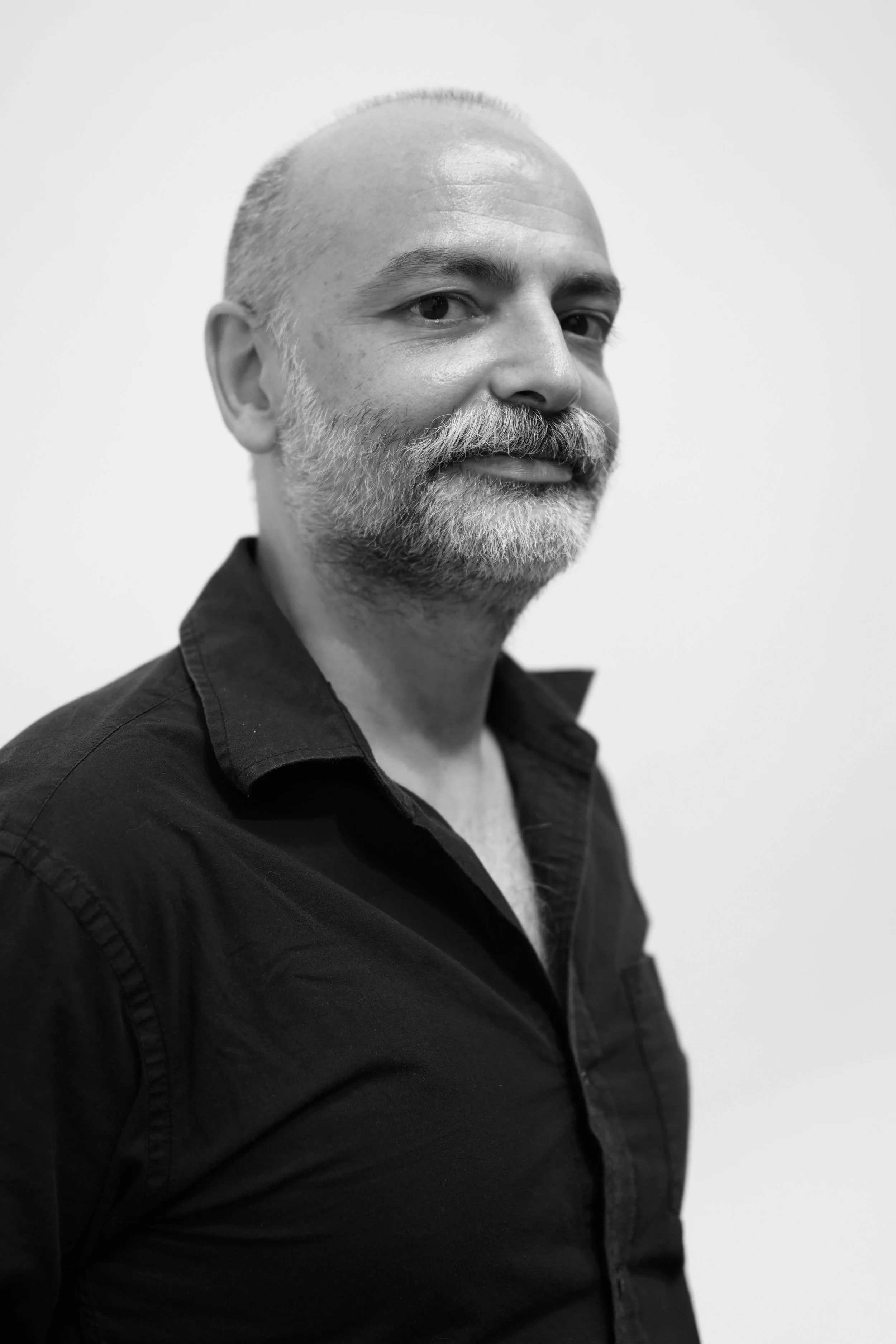 A black and white portrait of a middle-aged man with a beard, smiling slightly, wearing a dark shirt, against a plain light background.