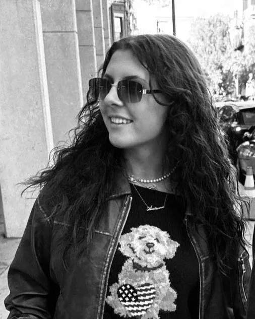 A woman with curly hair wearing sunglasses, a pearl necklace, a graphic sweater with a dog holding an American flag, and a leather jacket, standing on a city street.