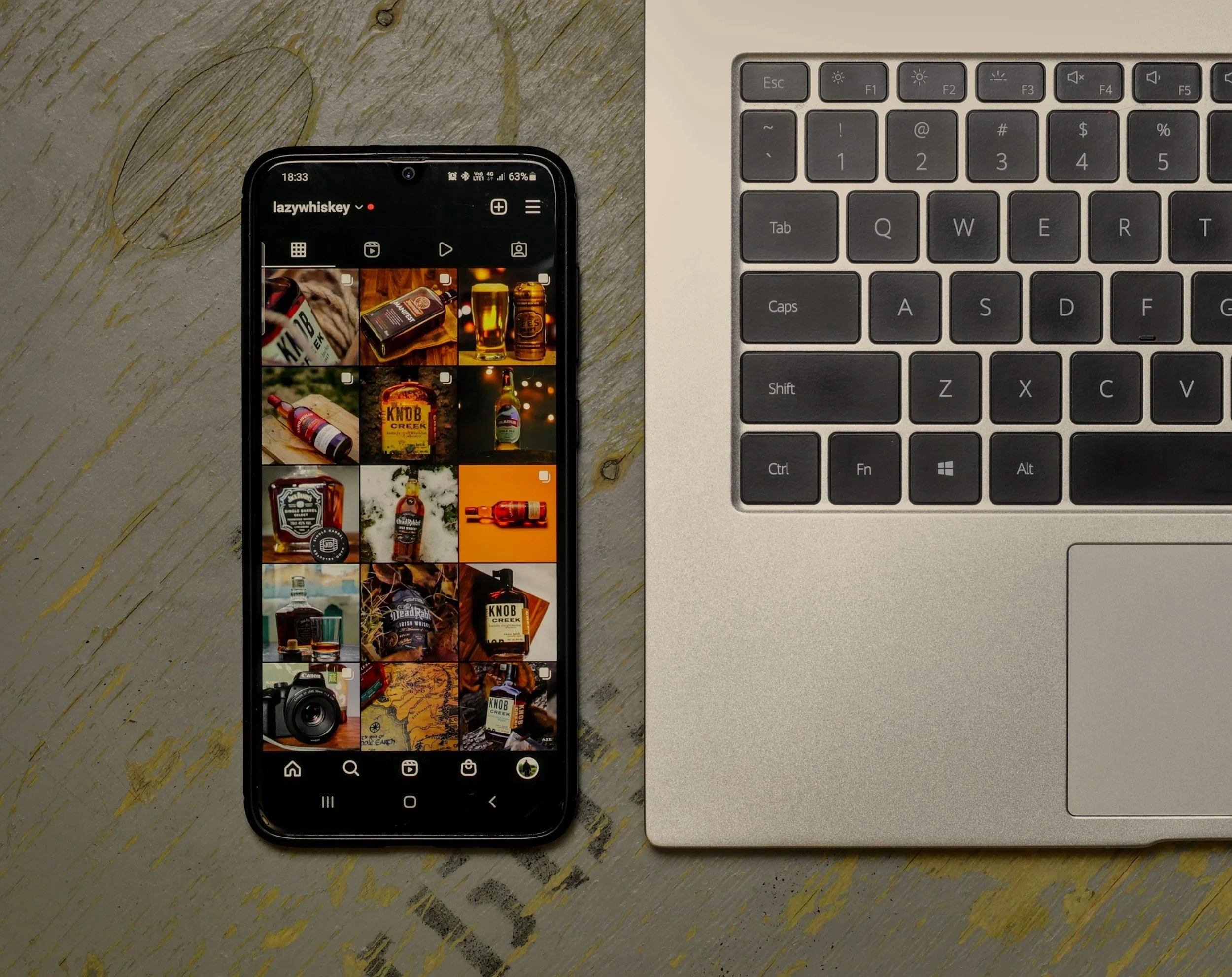 A smartphone displaying an Instagram profile with pictures of whiskey bottles, next to a silver laptop keyboard on a wooden desk.