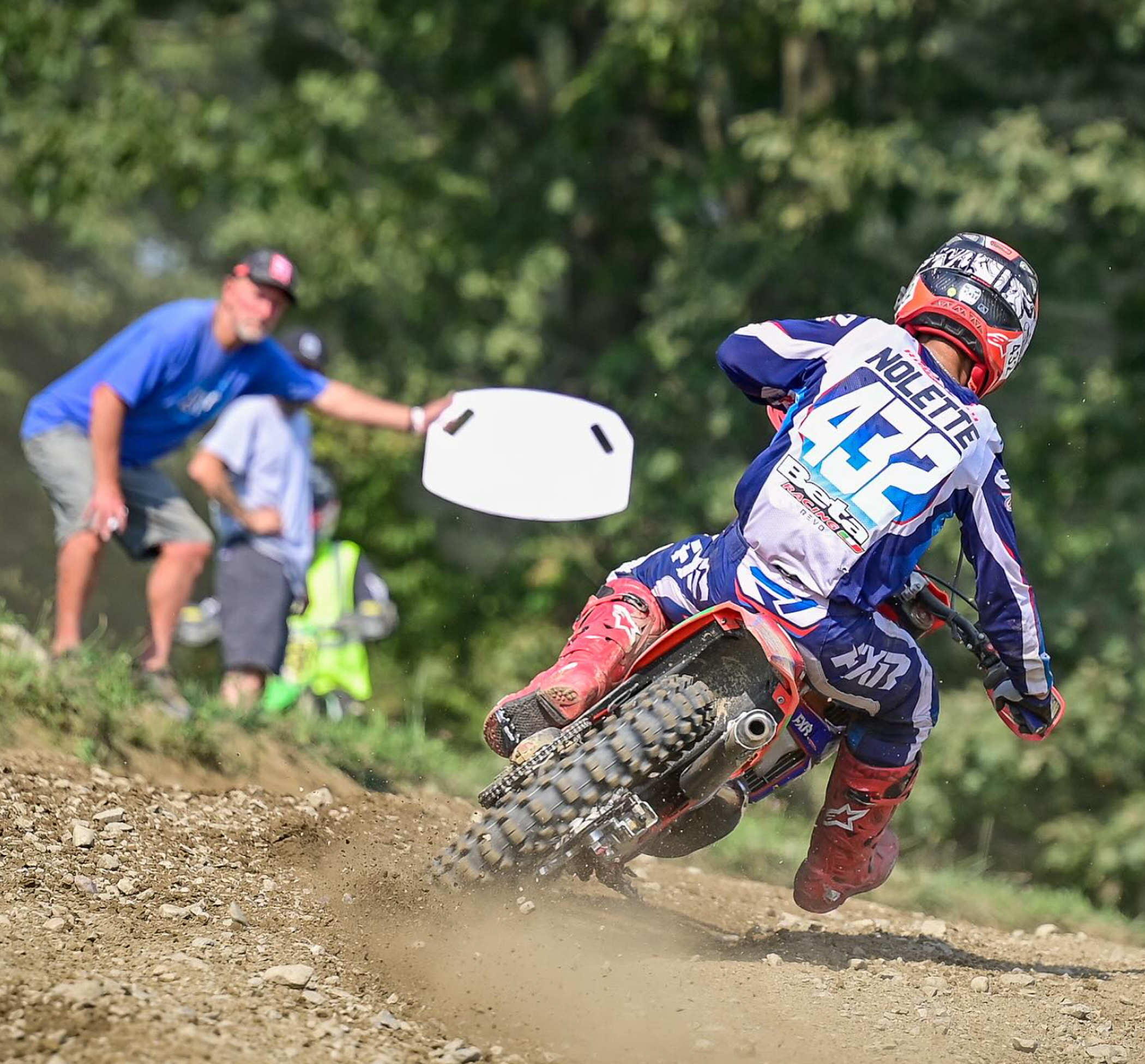 Motocross rider in blue gear and a red helmet racing on a dirt track, with spectators and trees in the background.