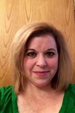 A woman with shoulder-length blonde hair, wearing a green top, standing in front of a wooden door.