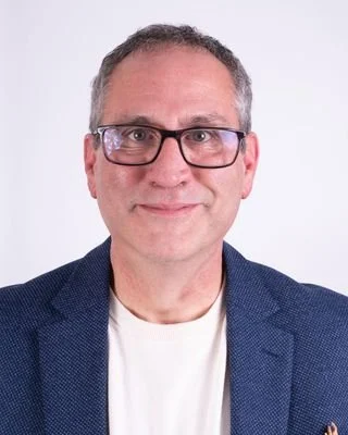 A middle-aged man with glasses, wearing a dark blue blazer and a white shirt, standing against a plain white background.