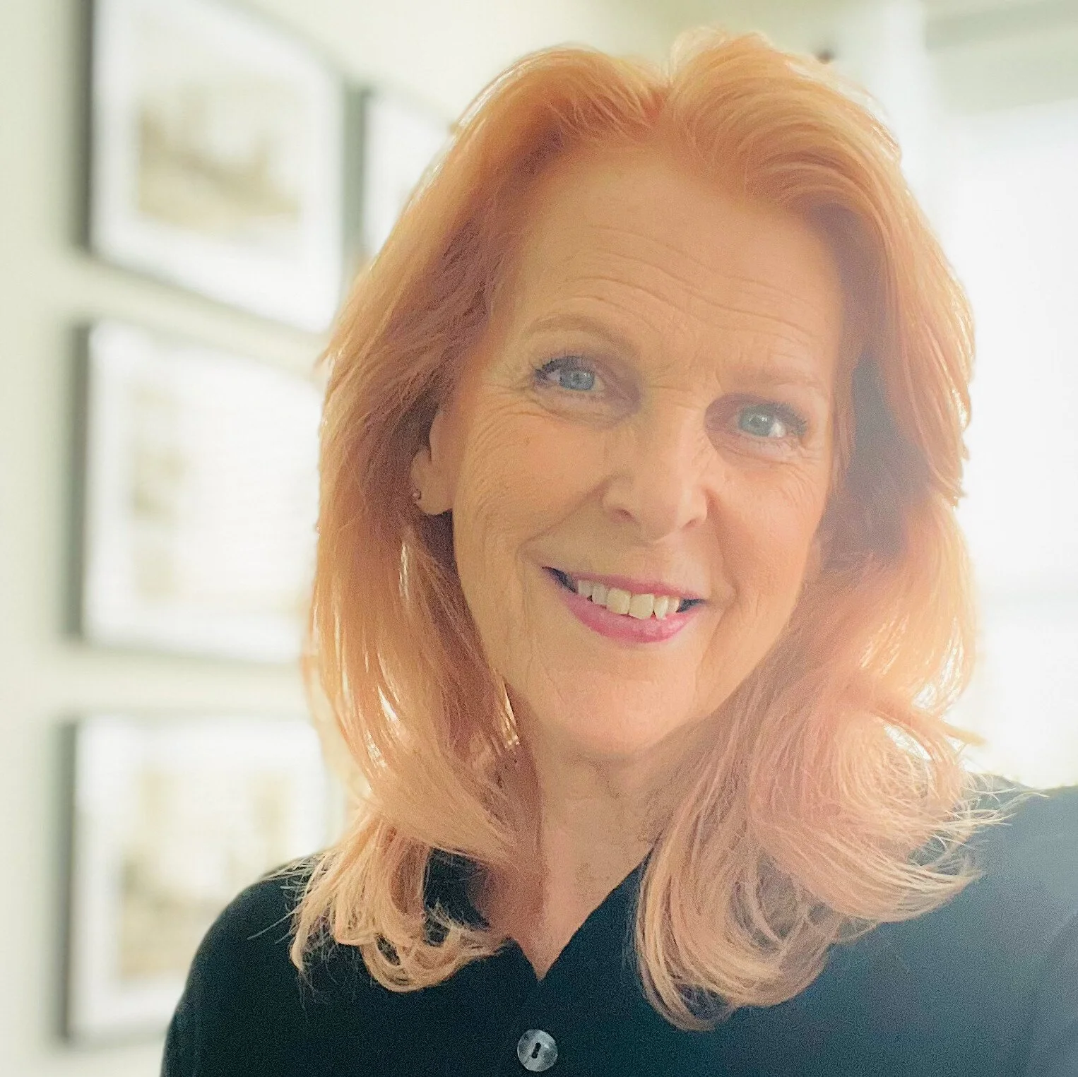 Close-up of a smiling woman with red hair and blue eyes, wearing a black top, standing in front of a window with sunlight coming through.