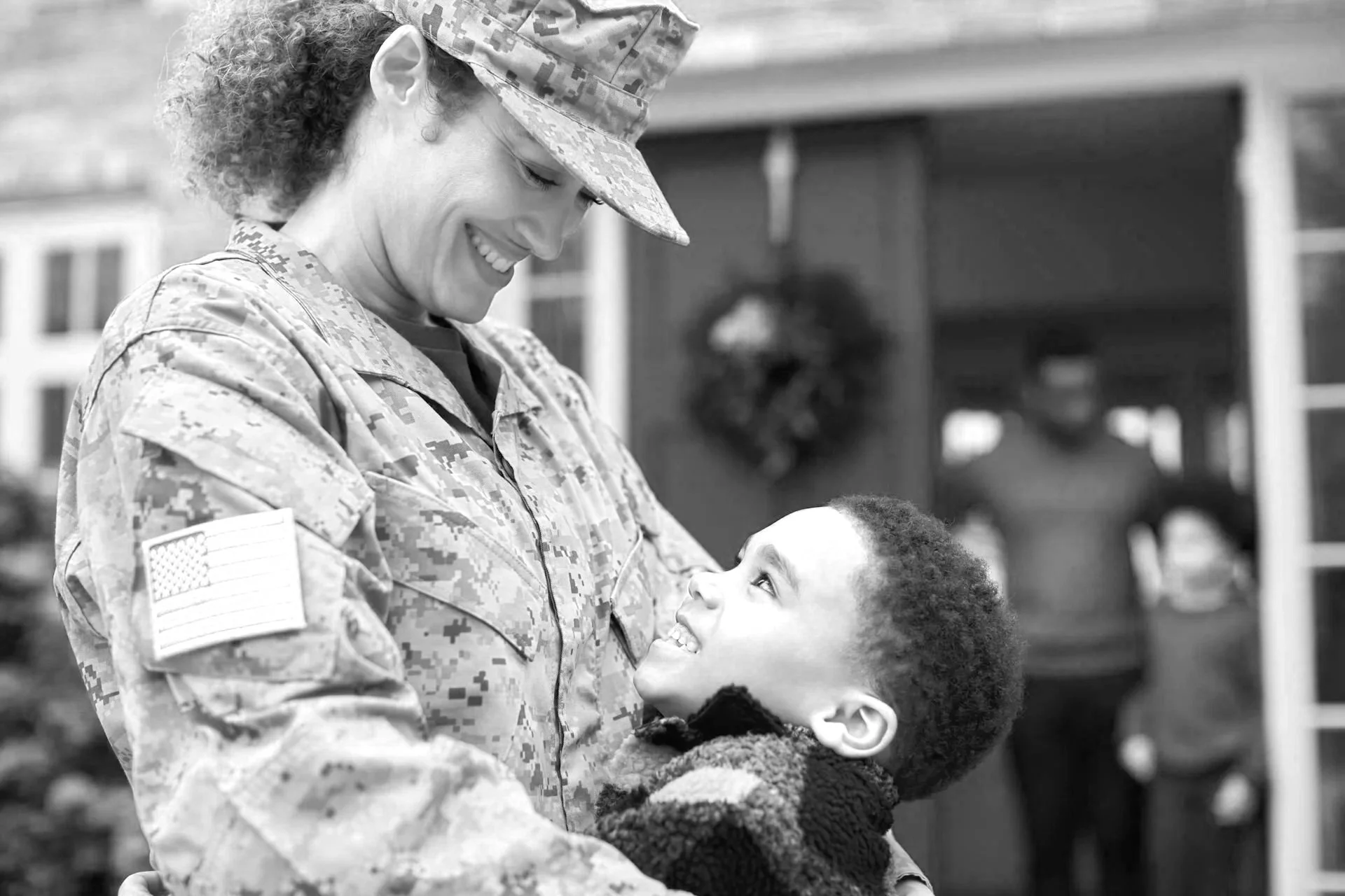 A female soldier in uniform holding a young boy, smiling and looking into each other's eyes outdoors.