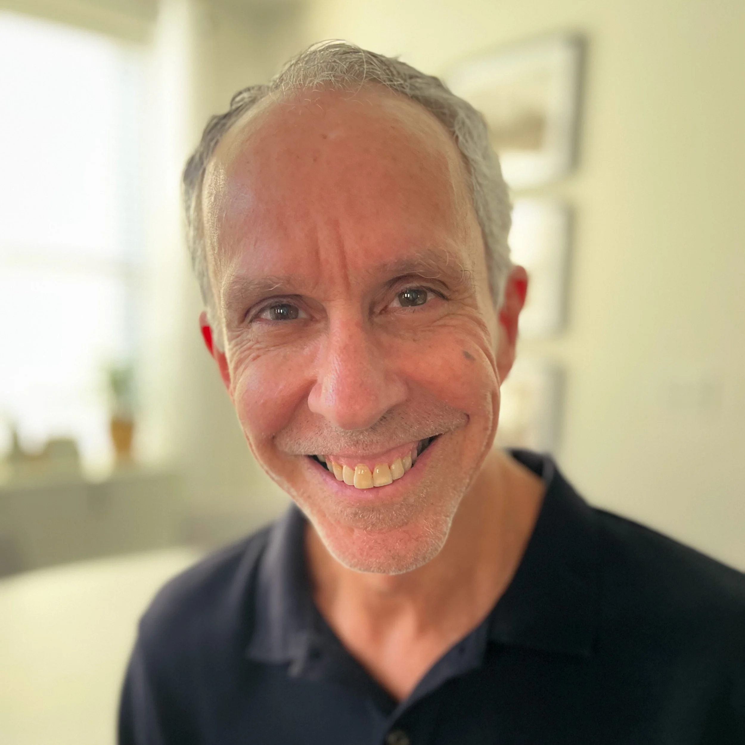 Close-up of smiling middle-aged man with short gray hair, wearing a black collared shirt, in a well-lit room with blurred background.