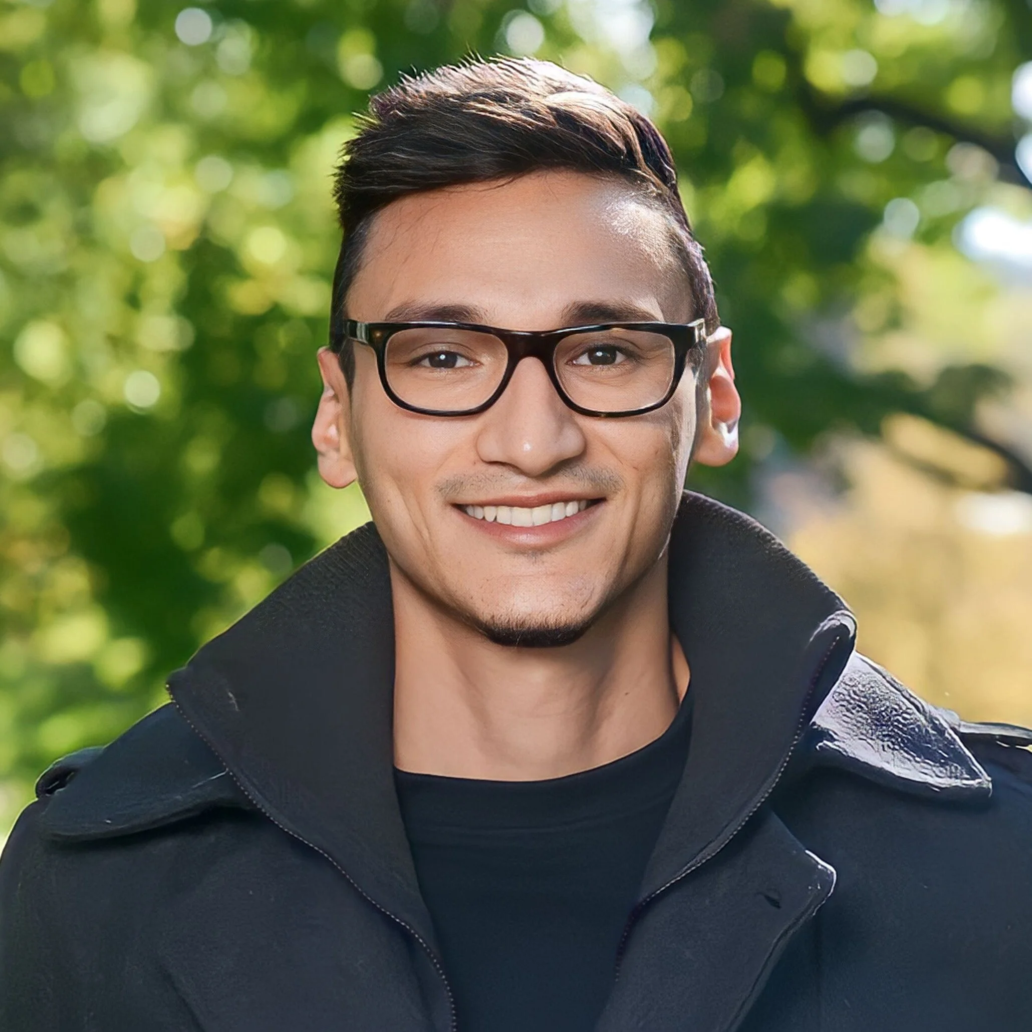 A young man with short dark hair, wearing black glasses, smiling, in an outdoor setting with green blurred foliage in the background.
