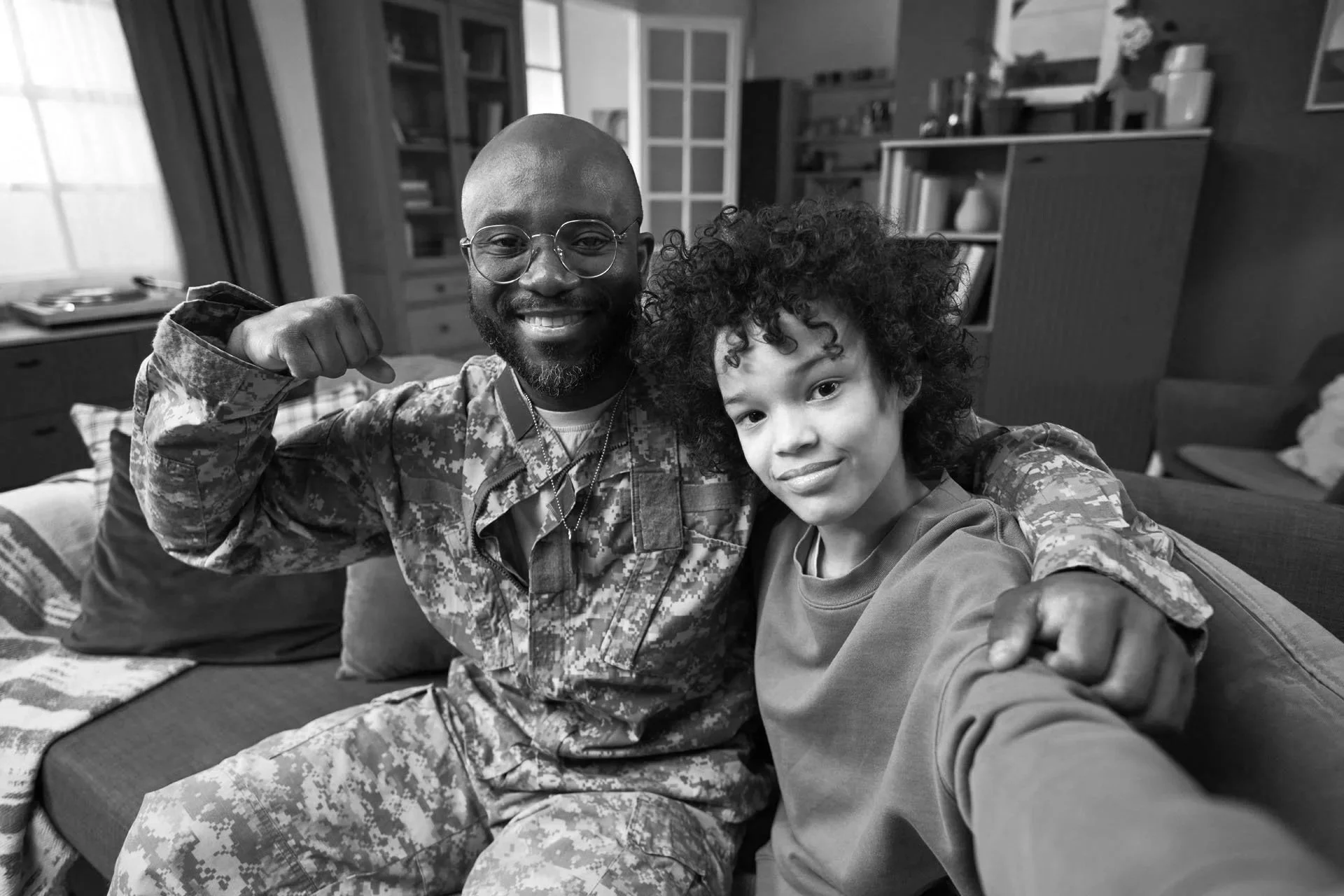 A man in military uniform and a young boy are sitting on a couch, smiling at the camera. The man is flexing his arm, and the boy is taking a selfie in a cozy living room.