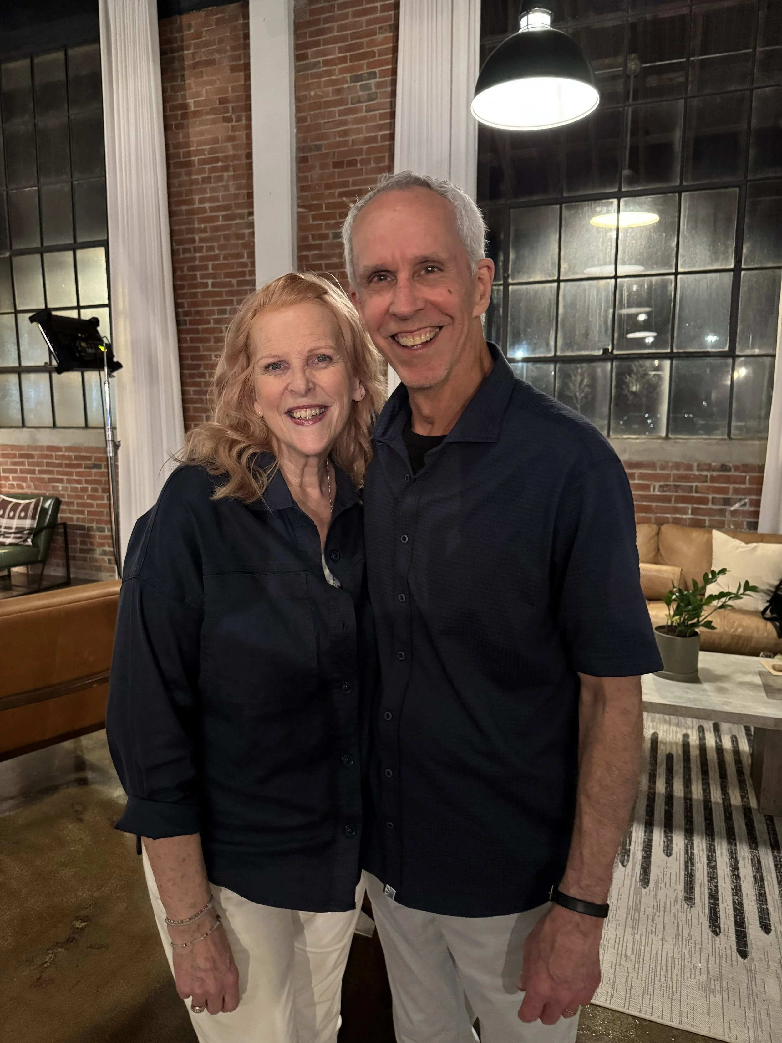 A smiling elderly woman and a smiling middle-aged man standing close together in a warmly lit, modern living room with brick walls, large industrial-style windows, a hanging lamp, a tan sofa, and houseplants.