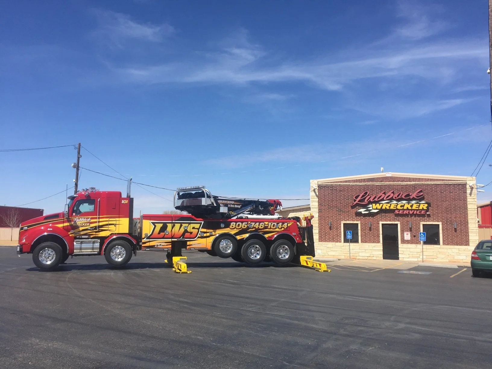 A red tow truck with yellow and black graphics parked in front of a building with a sign that reads 'Lubbock Wrecker Service.' The truck has a large crane and yellow wheel chocks, with a visible phone number on the side. The parking lot has blue handicap spaces and a green car partially visible on the right, under a clear blue sky.