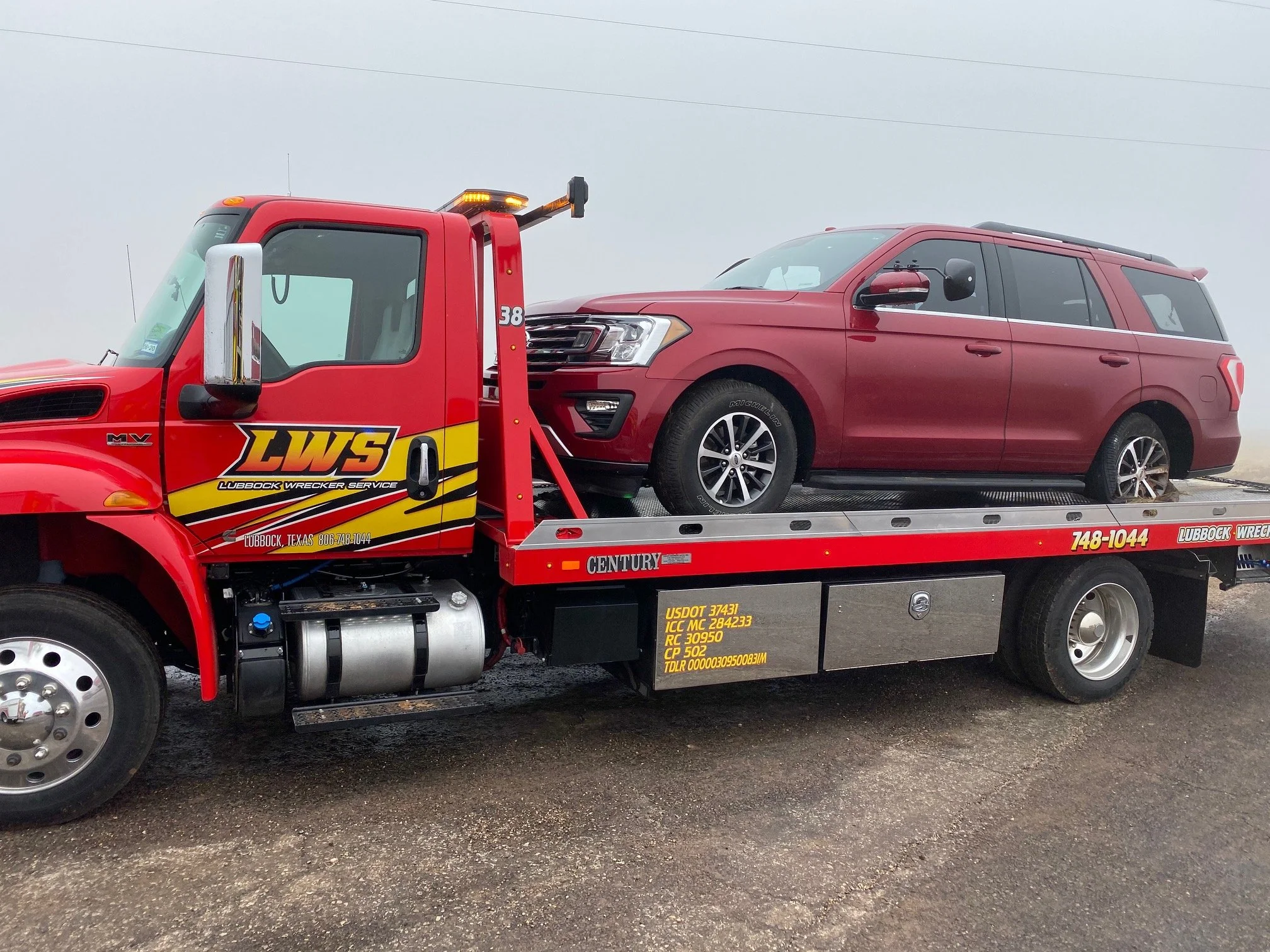 Red tow truck labeled "LWS Lubbock Wrecker Service" carrying a red SUV with a visible side mirror and a long flatbed, parked on a gravel surface with an overcast sky.