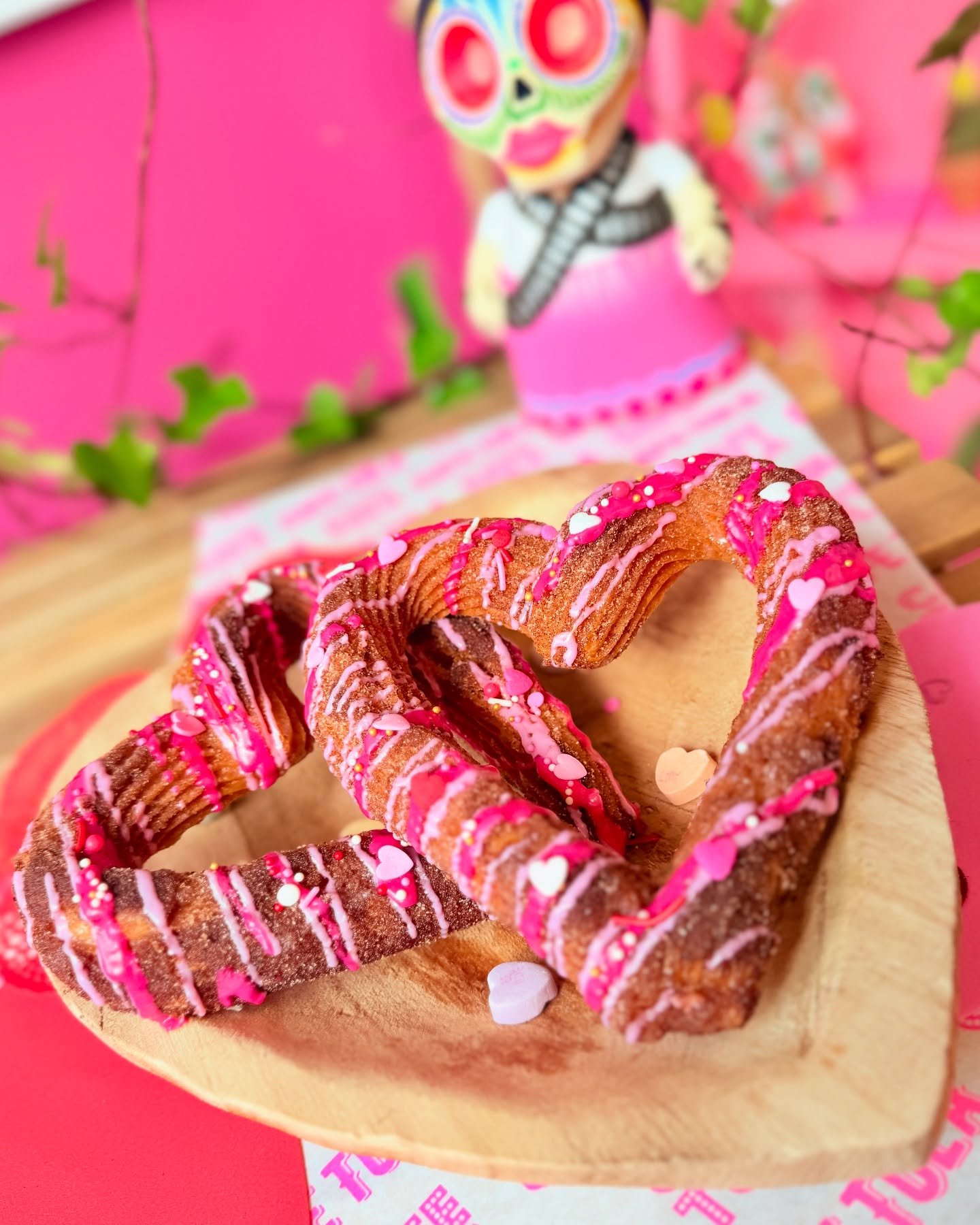 Heart-shaped cookies decorated with pink icing and sprinkles on a wooden plate, with a colorful skeleton figure in the background.