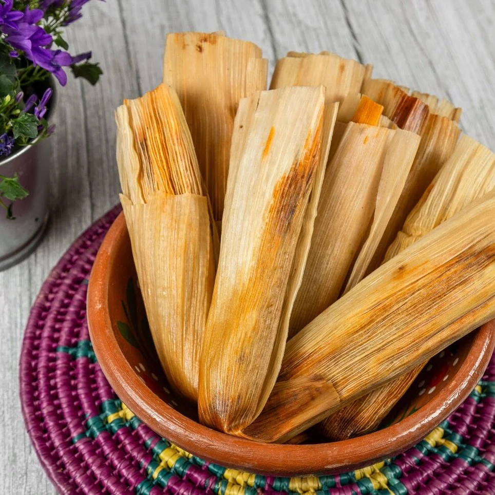Tamales wrapped in corn husks in a clay bowl on a woven pink and purple placemat.