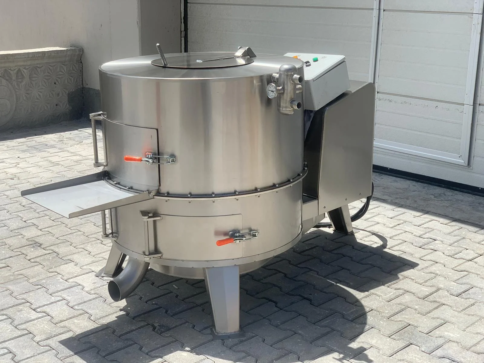 Industrial stainless steel machine with a large cylindrical chamber, control panel, and a tray attached to the side, placed on a paved outdoor area.