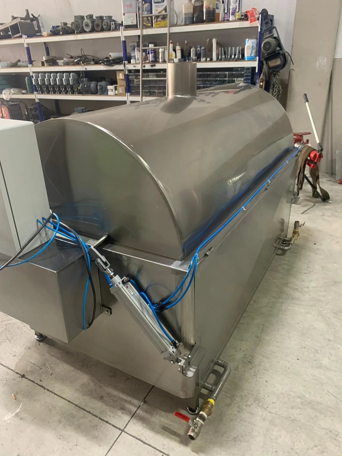 Stainless steel industrial machine in a workshop, with pipes, tubes, and control panel, used for manufacturing or processing.