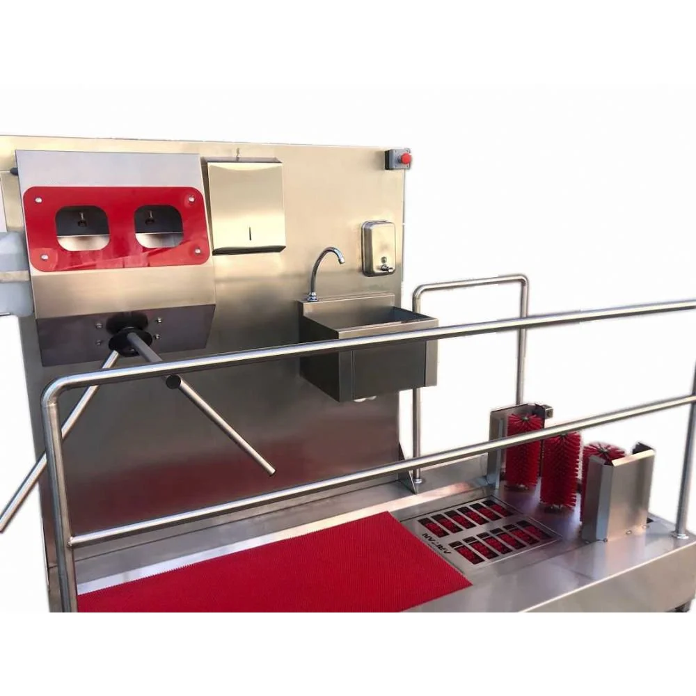 A stainless steel industrial cleaning station with a sink, paper towel dispenser, hand sanitizer, and a red mat with red brushes, surrounded by metal safety rails.