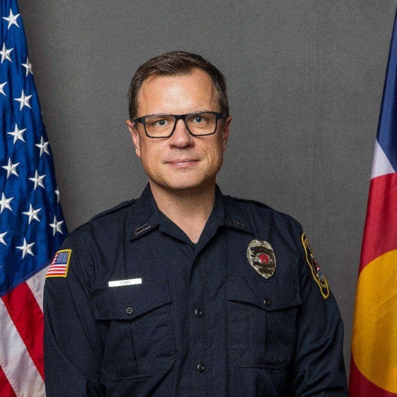 A man in a police uniform standing in front of American and Colorado flags.