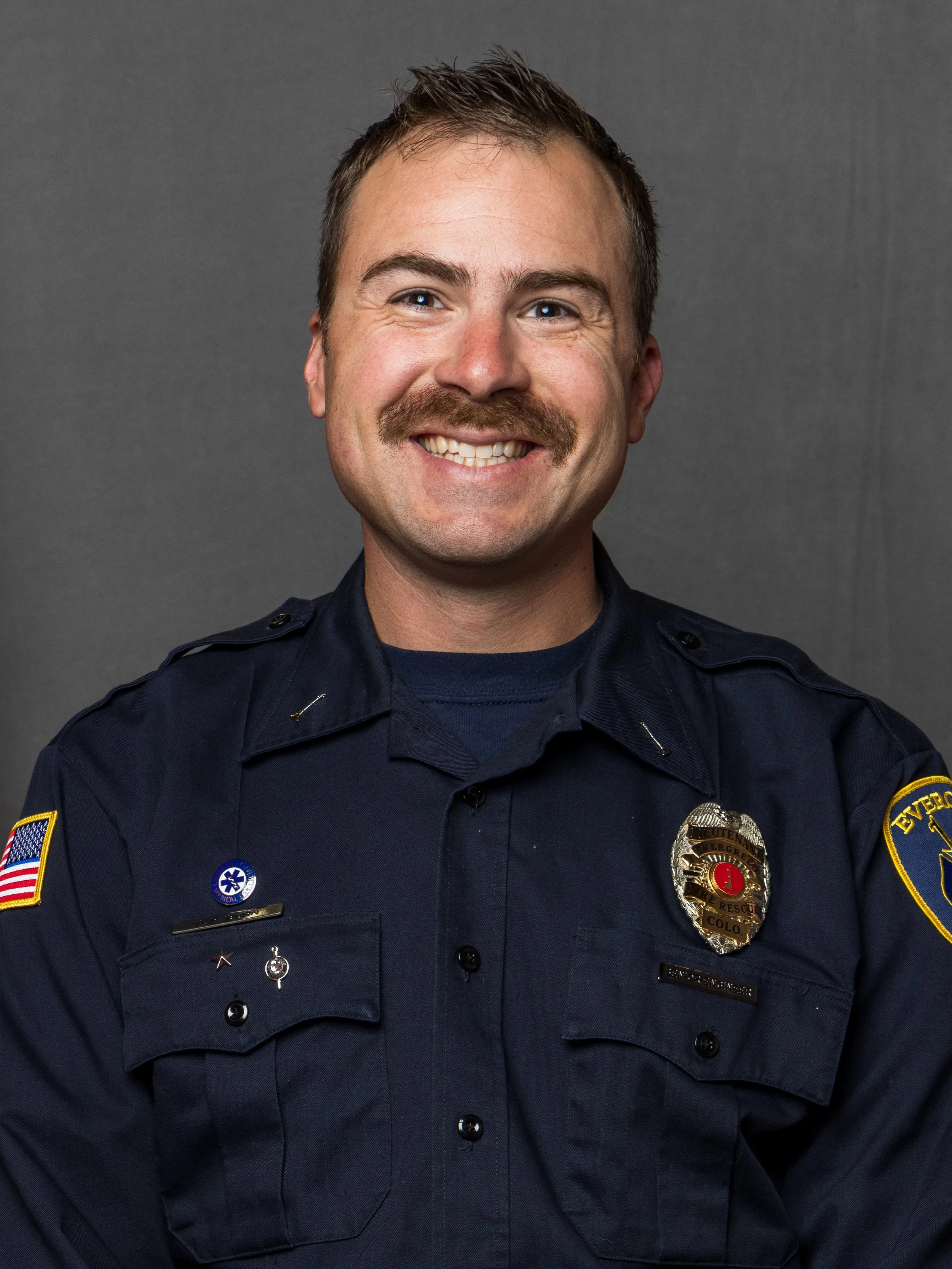 A smiling male police officer in uniform with badges and patches, posing against a plain dark background.