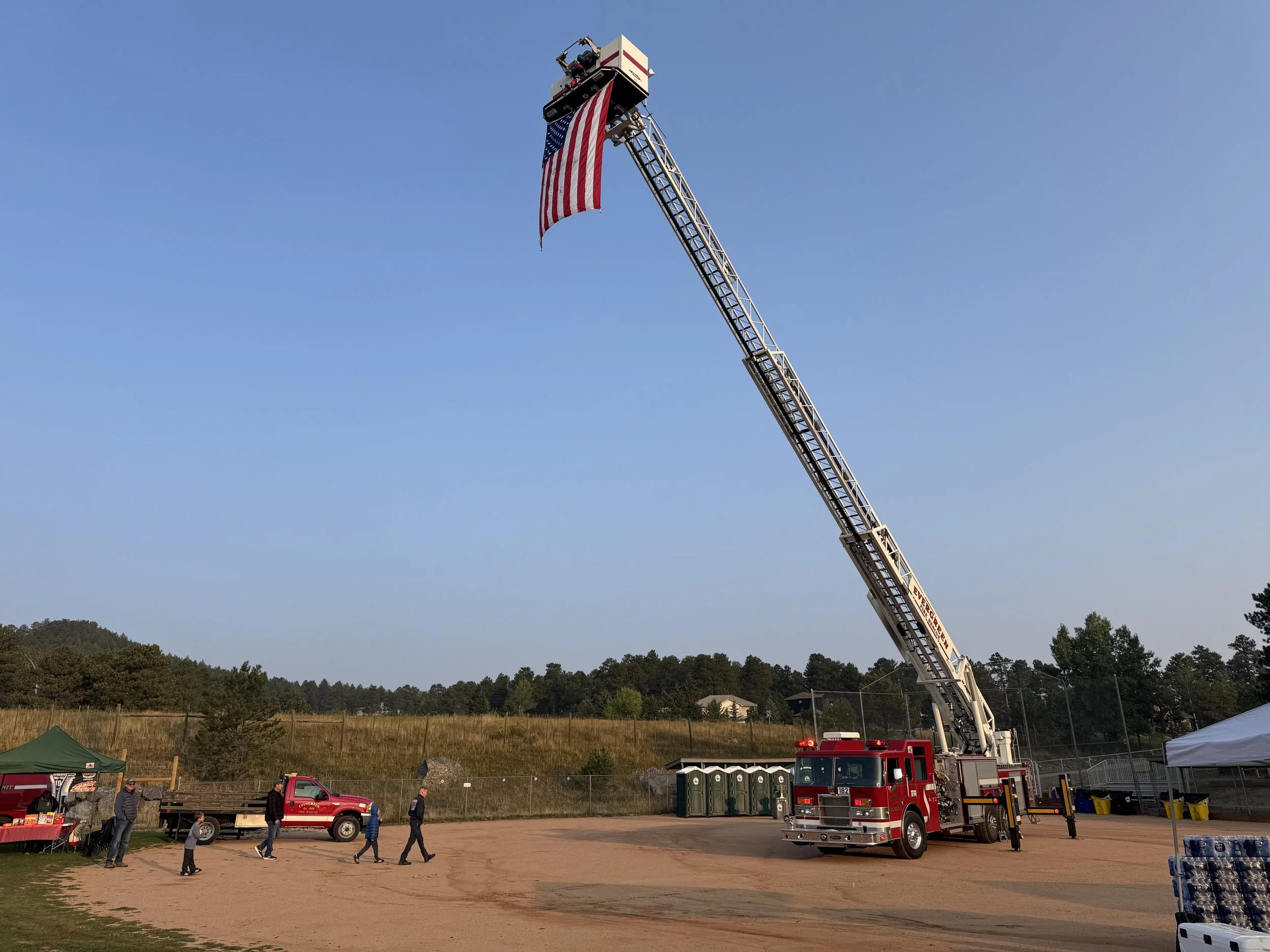 A fire truck with an extended ladder holding an American flag in a field with people walking nearby and tents at a festival or event.