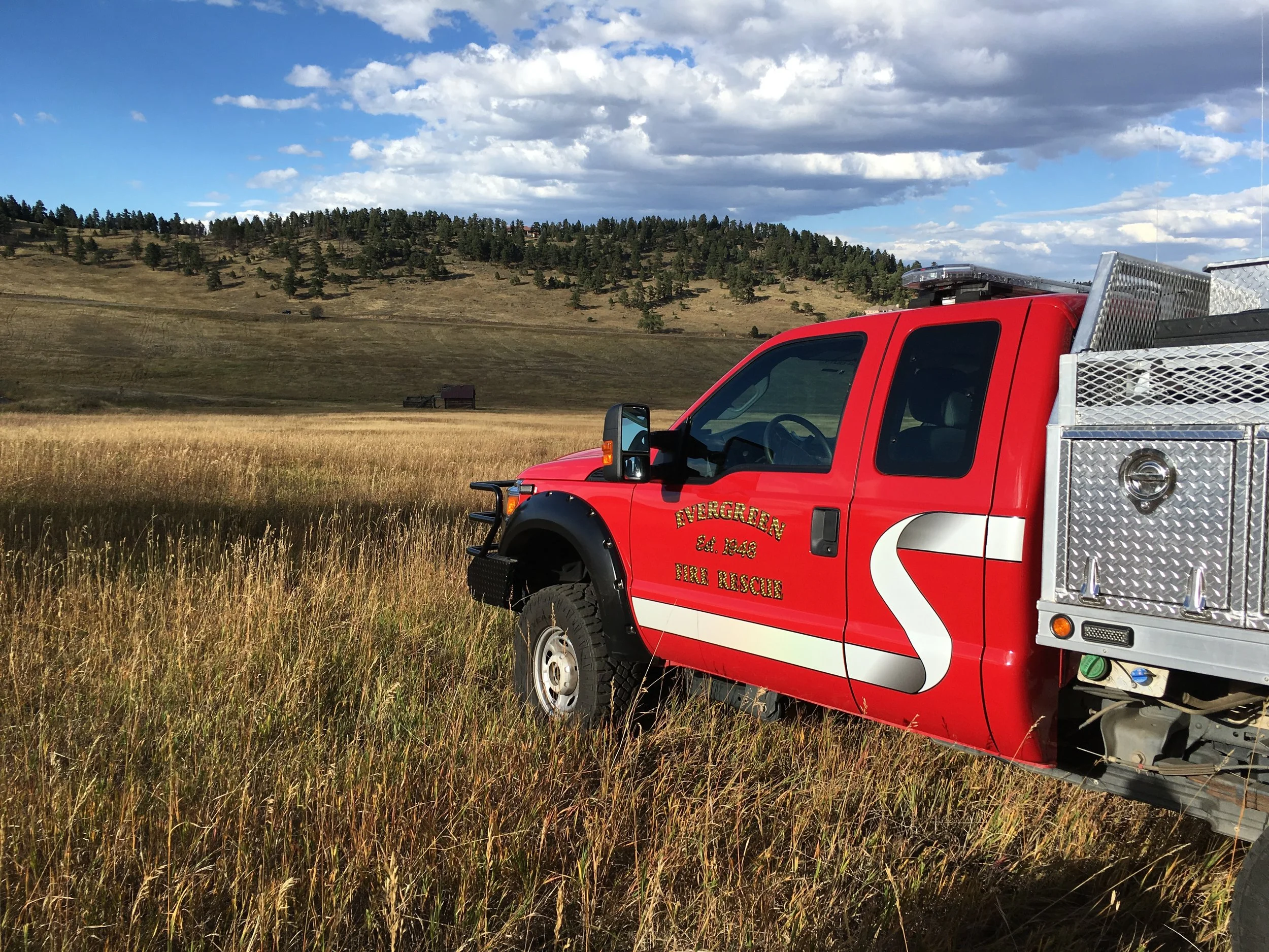 A red fire rescue vehicle parked in a grassy field with a hill covered in trees in the background under a partly cloudy sky.