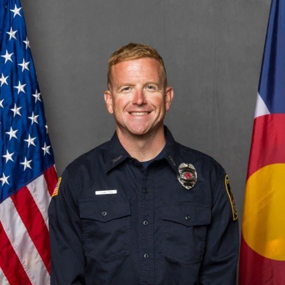 A smiling police officer in uniform standing in front of the American and Colorado flags.