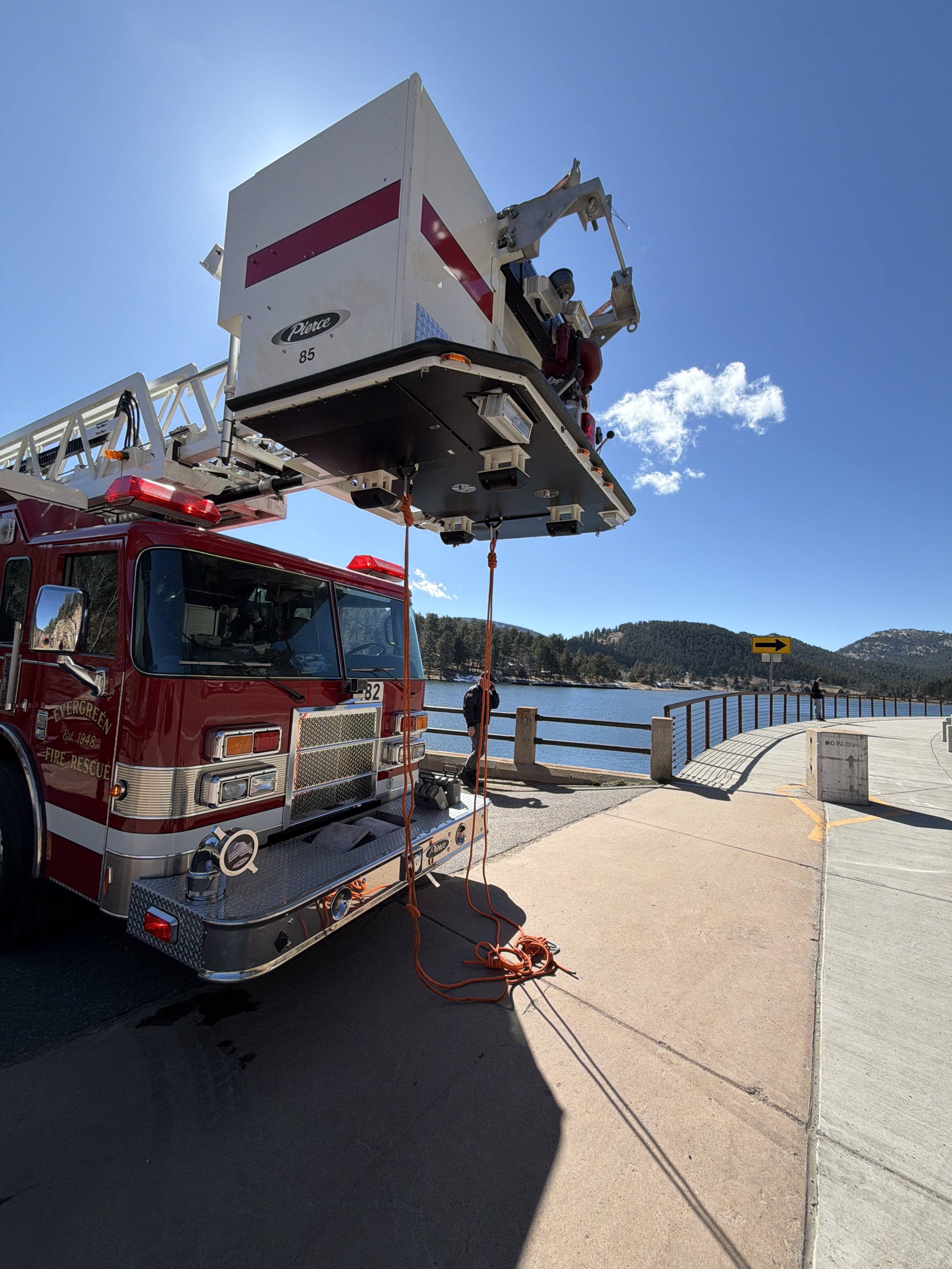 Fire truck with an elevated platform, a firefighter standing nearby, scenic water and mountain background with clear blue sky.