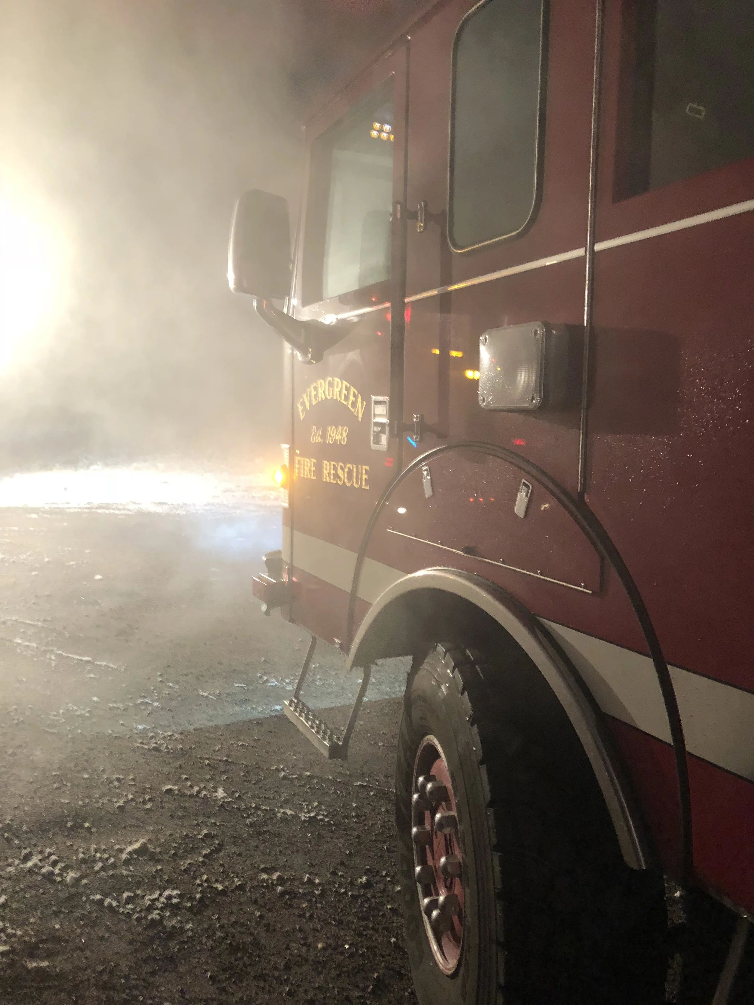 A fire rescue truck at night with foggy conditions, showing part of the truck's side with the words 'Evergreen Fire Rescue' and the establishment year 1948, along with a side mirror, a window, and a wheel.