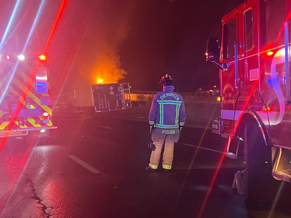 Firefighter standing on the road at night, looking at a vehicle burning with flames and smoke, surrounded by emergency vehicles with flashing lights.