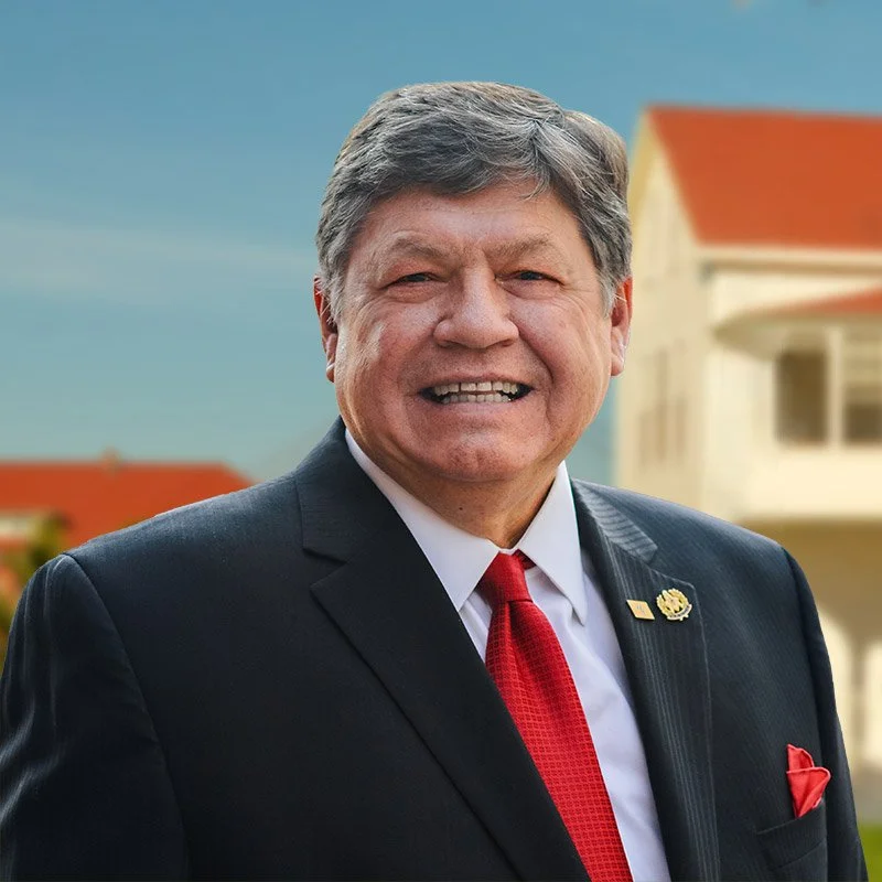 A smiling middle-aged man in a dark suit with a red tie and white shirt, standing outdoors with a blurred house in the background.