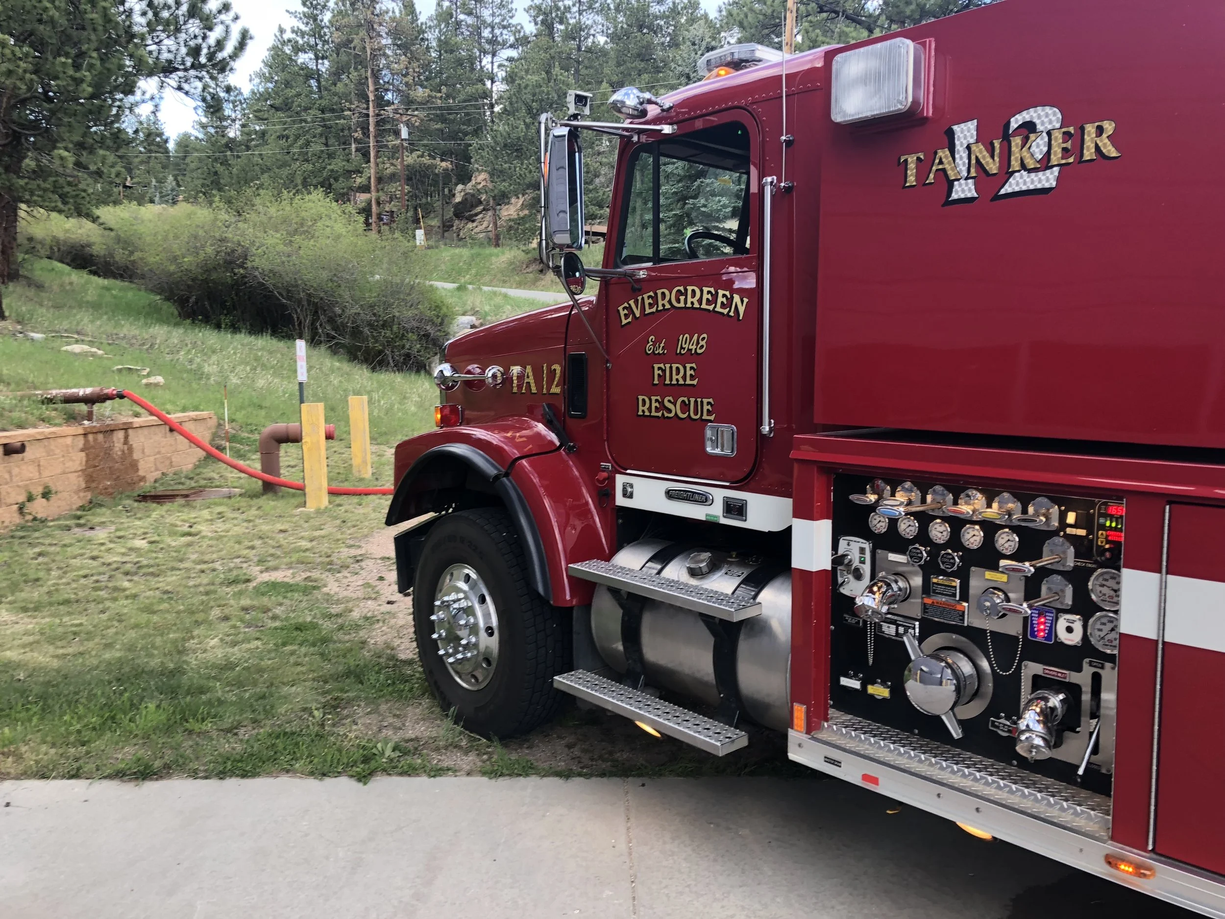 A red fire rescue truck with the words 'Evergreen Fire Rescue' and 'Tanker 12' on the side, parked on a grassy area near trees, with firefighting equipment and gauges visible.
