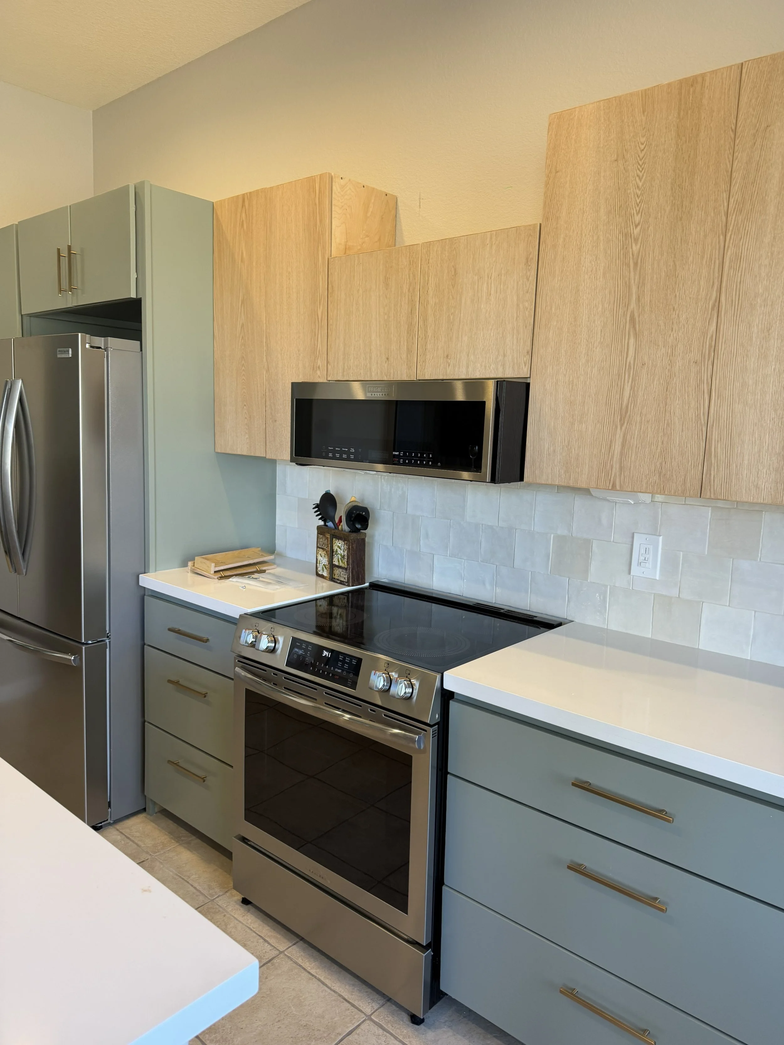 A modern kitchen with gray and wood cabinets, stainless steel appliances including a refrigerator, microwave, and oven, and a tiled backsplash.