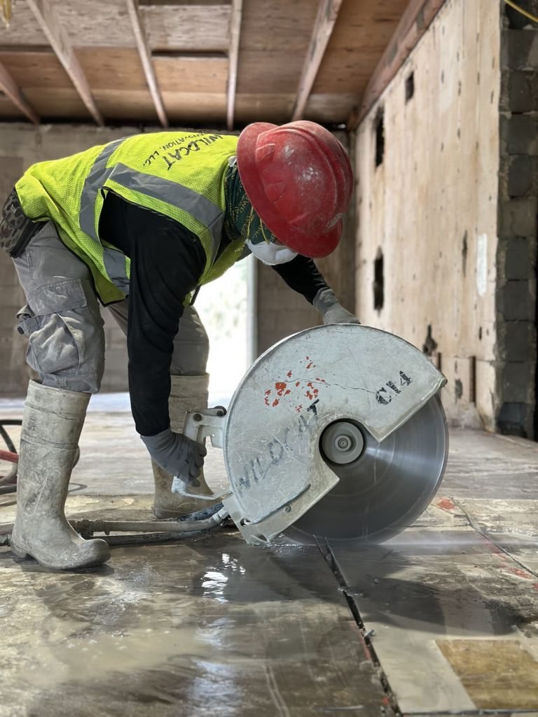 Construction worker in a red hard hat, yellow safety vest, and gray gloves cuts a tile floor with a large electric saw inside a building under construction.