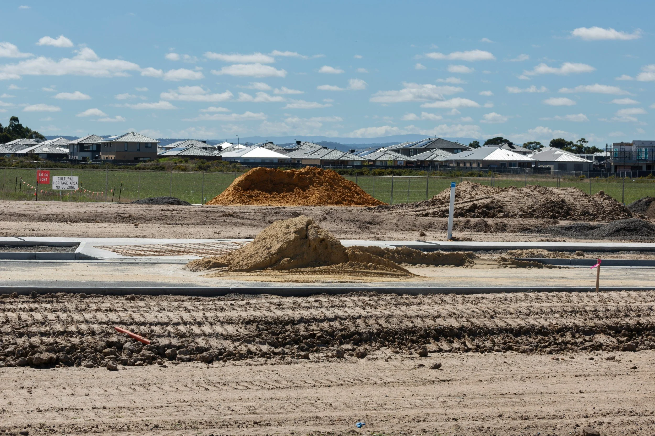 View of a construction site with piles of dirt and construction materials, with residential houses in the background under a partly cloudy sky.