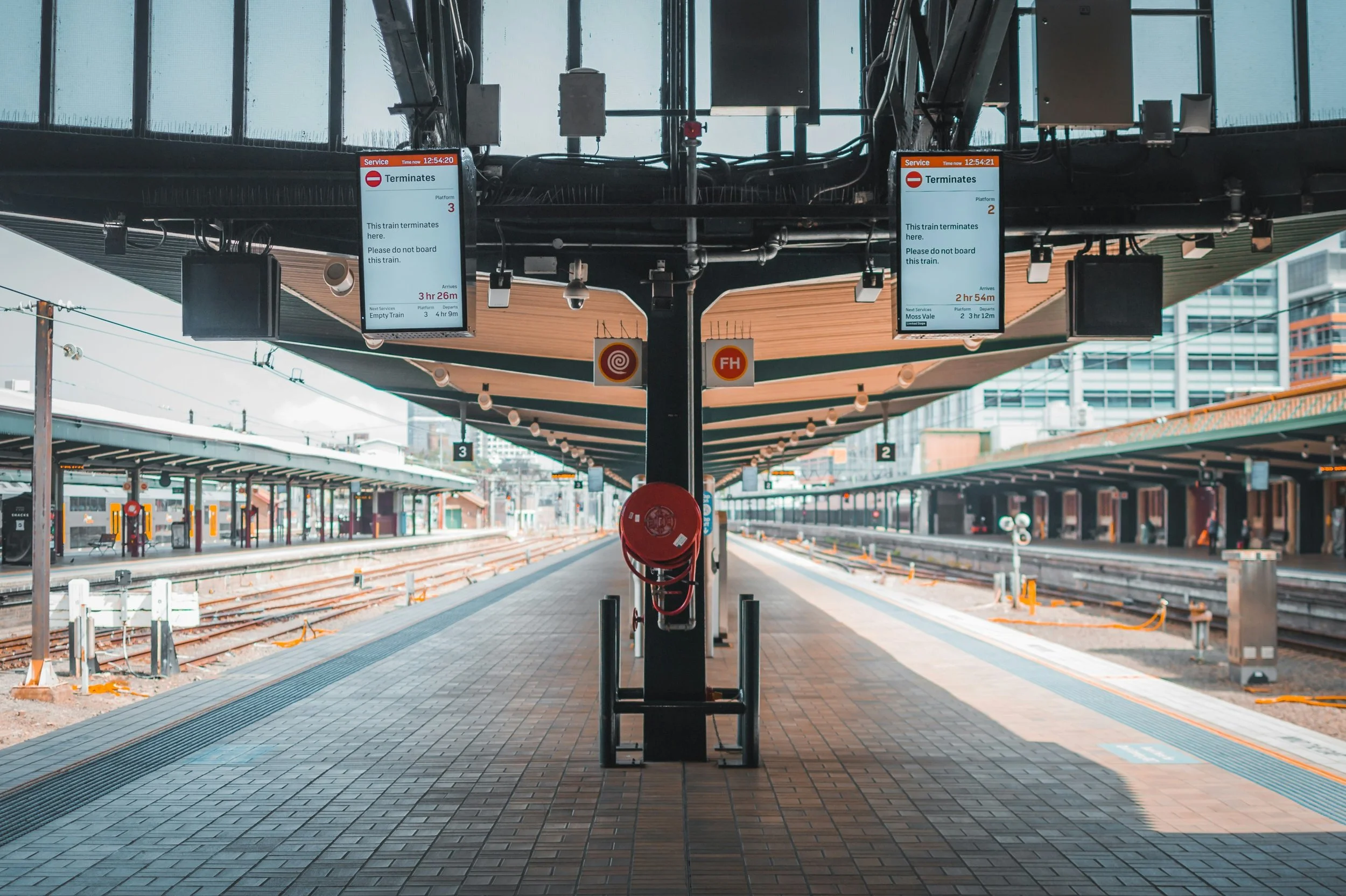 Empty train station platform with digital signs indicating train termination, platforms 2 and 3, with some buildings and tracks in the background.