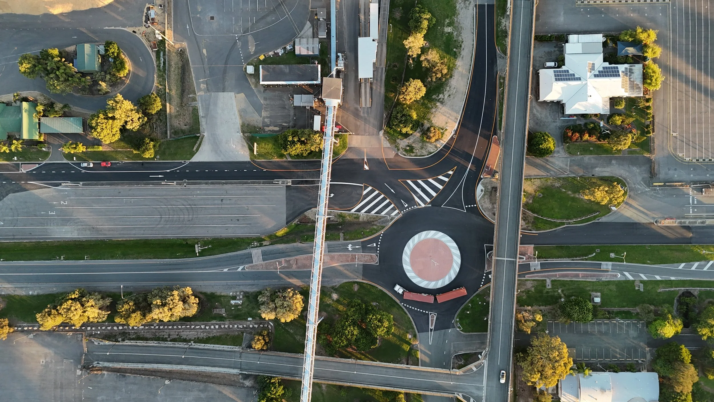 An aerial view of a city intersection with roads, parking lots, and buildings, including a white house with solar panels, surrounded by trees.