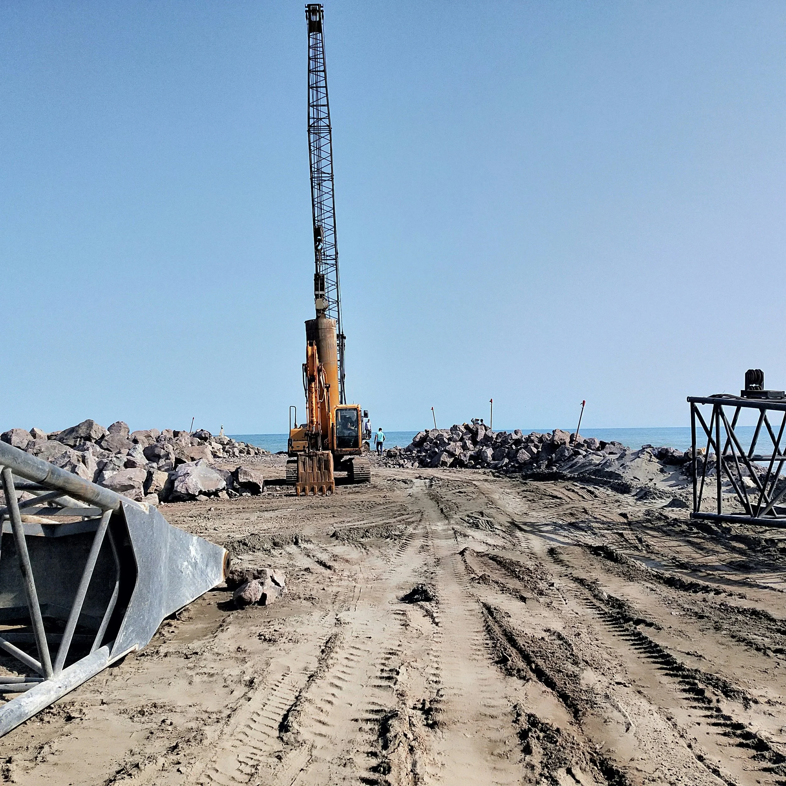 Construction site by the beach with an excavator working on a rock barrier, metal railings, and clear blue sky.