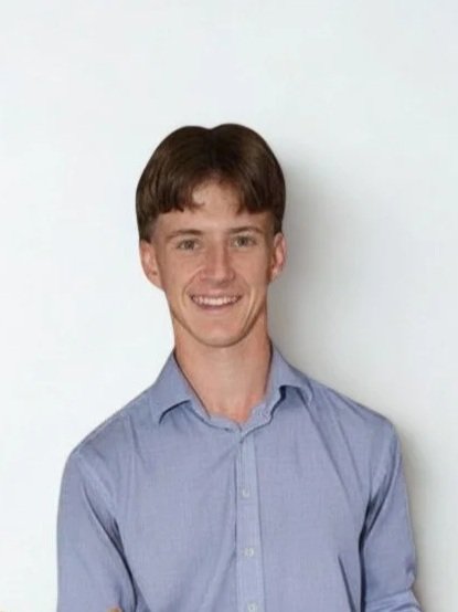 A young man with brown hair smiling, wearing a light blue button-up shirt, standing against a plain white wall.