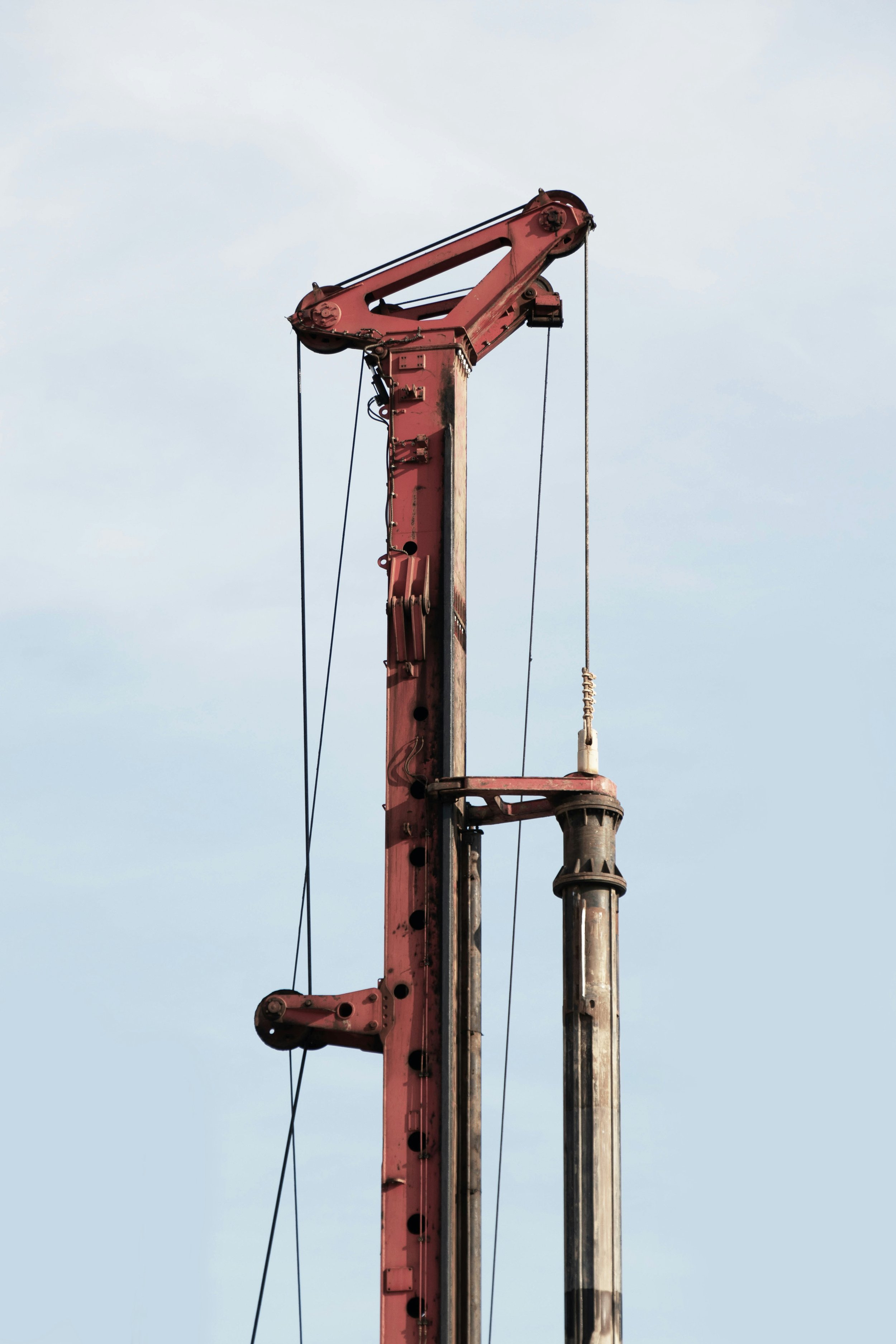 A tall, red and metallic crane structure against a cloudy light blue sky, with visible cables and pulleys.