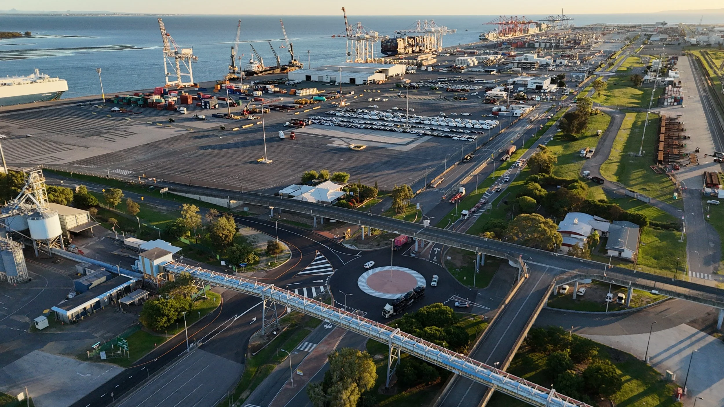 An aerial view of a port with dock cranes, cargo containers, and shipping vessels by the water, with roads, parking lots, and green areas in the foreground.