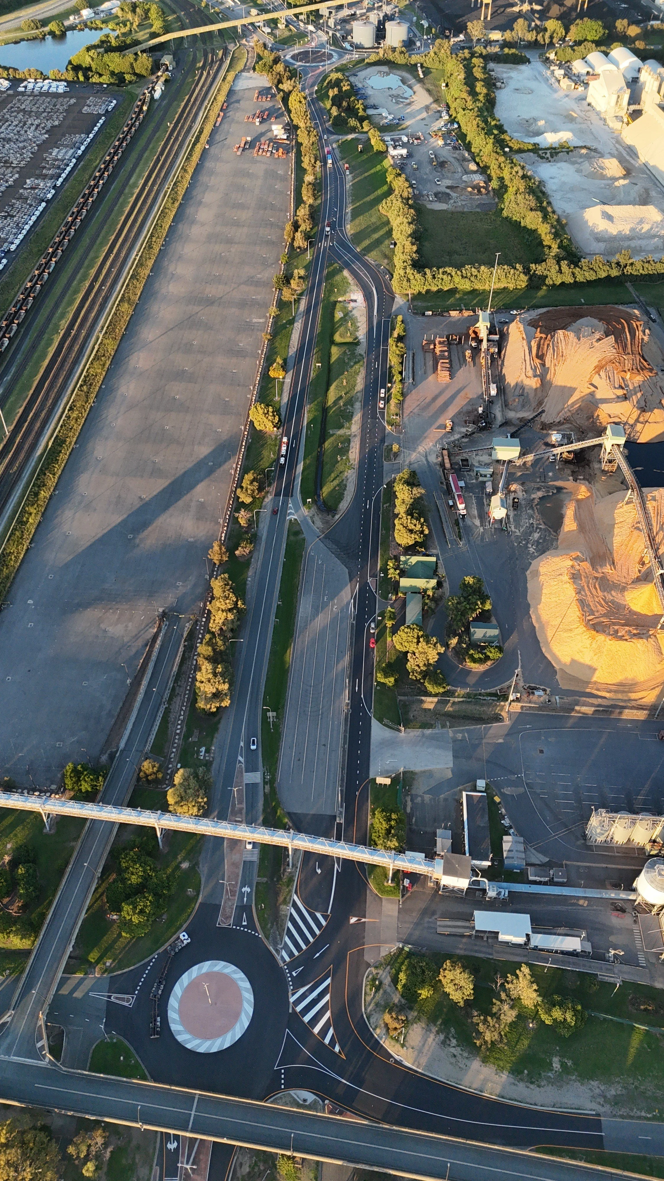 An aerial view of a multi-lane road with roundabouts, green areas, construction sites with sandpiles, parking lots, and industrial buildings surrounded by trees.