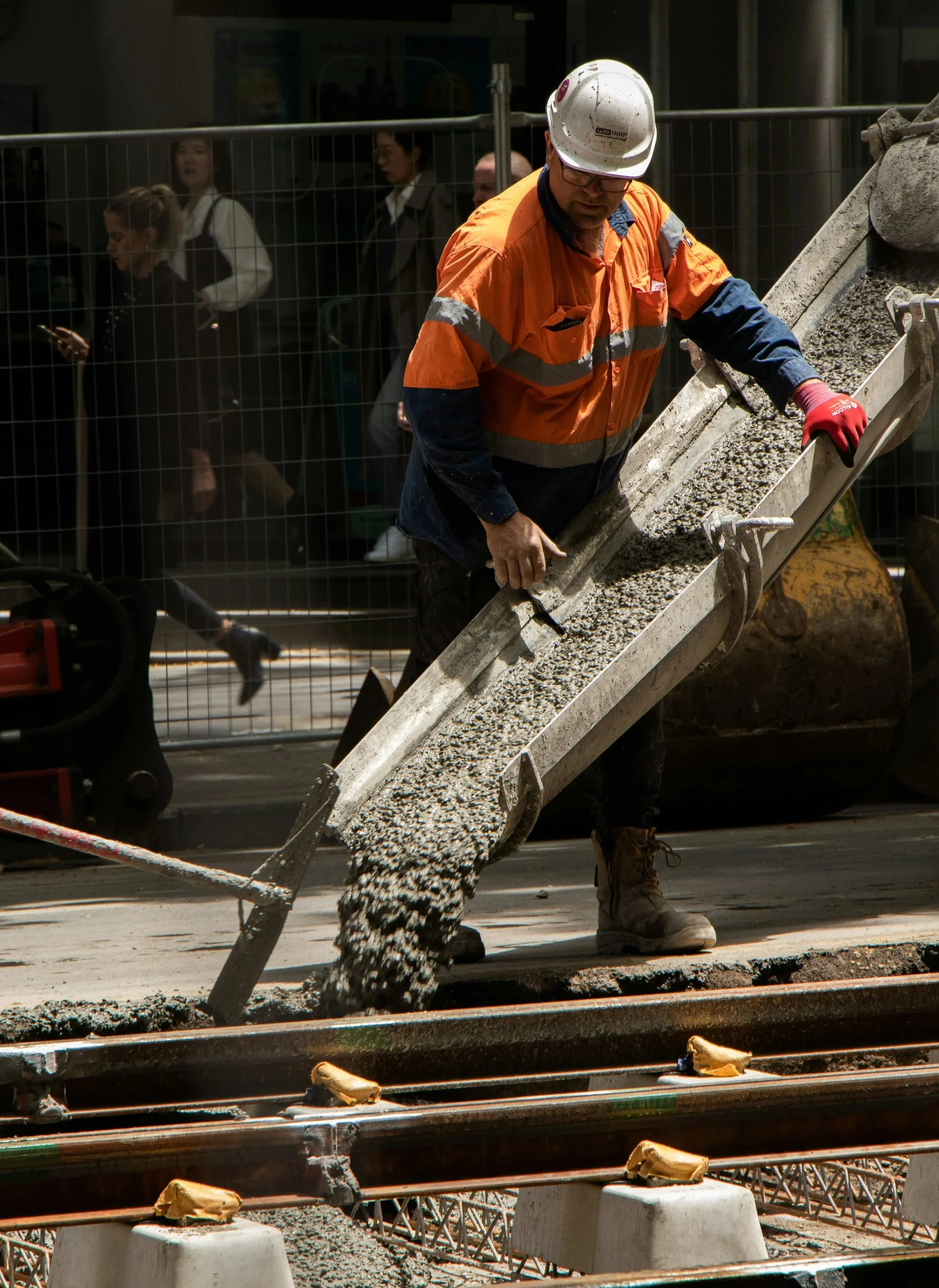 A construction worker wearing a white hard hat, orange safety vest, and red gloves is pouring wet concrete from a metal chute onto a surface. There are onlookers behind a safety barrier in the background.
