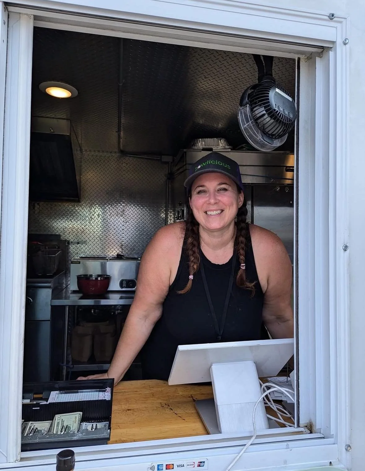 Smiling woman with braided hair and a black tank top working at a food truck window, wearing a black cap with green text, inside a stainless steel kitchen.